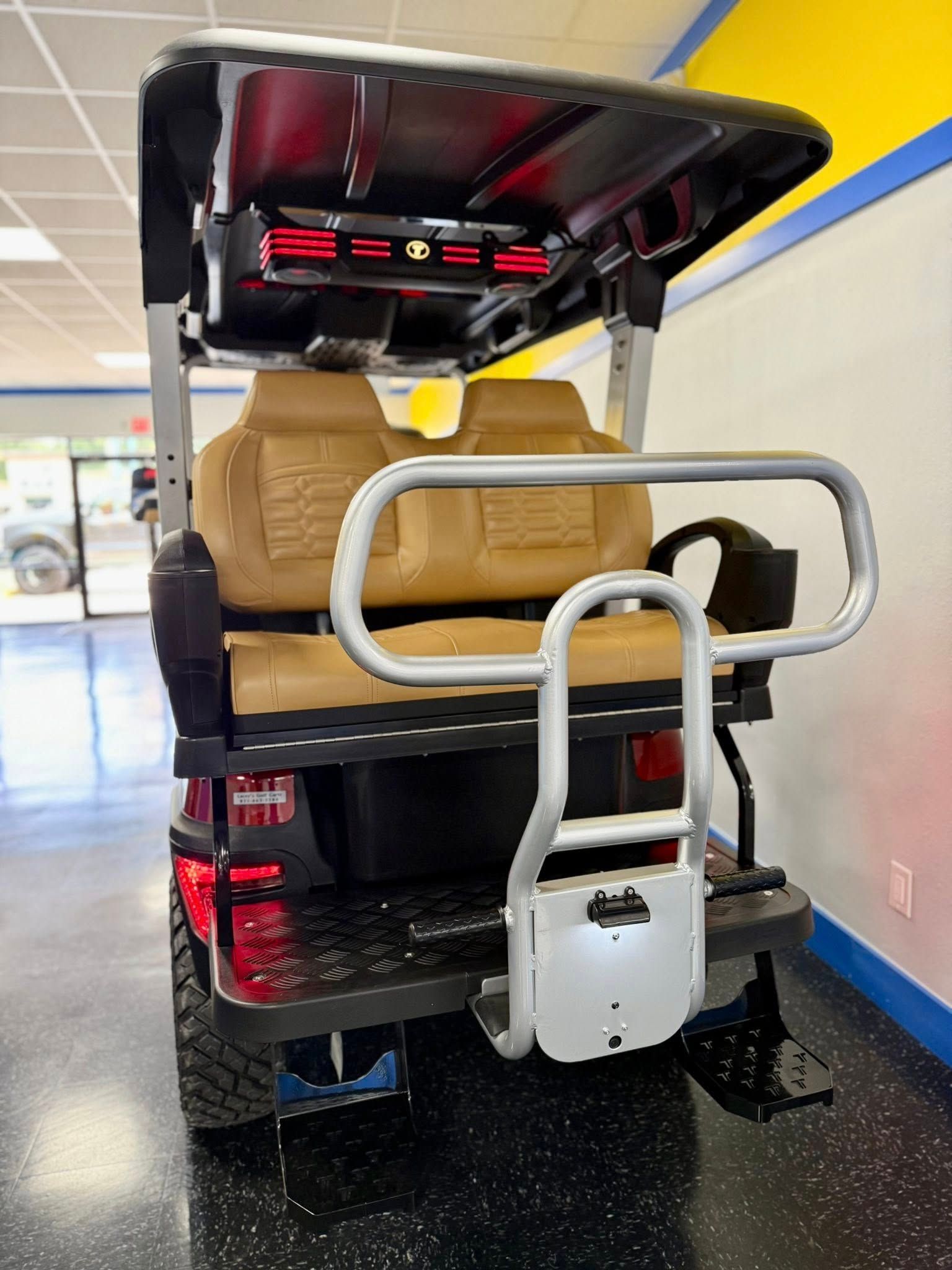 Rear view of a tan and black golf cart, with a rear-facing seat, in a showroom with blue and yellow accents.