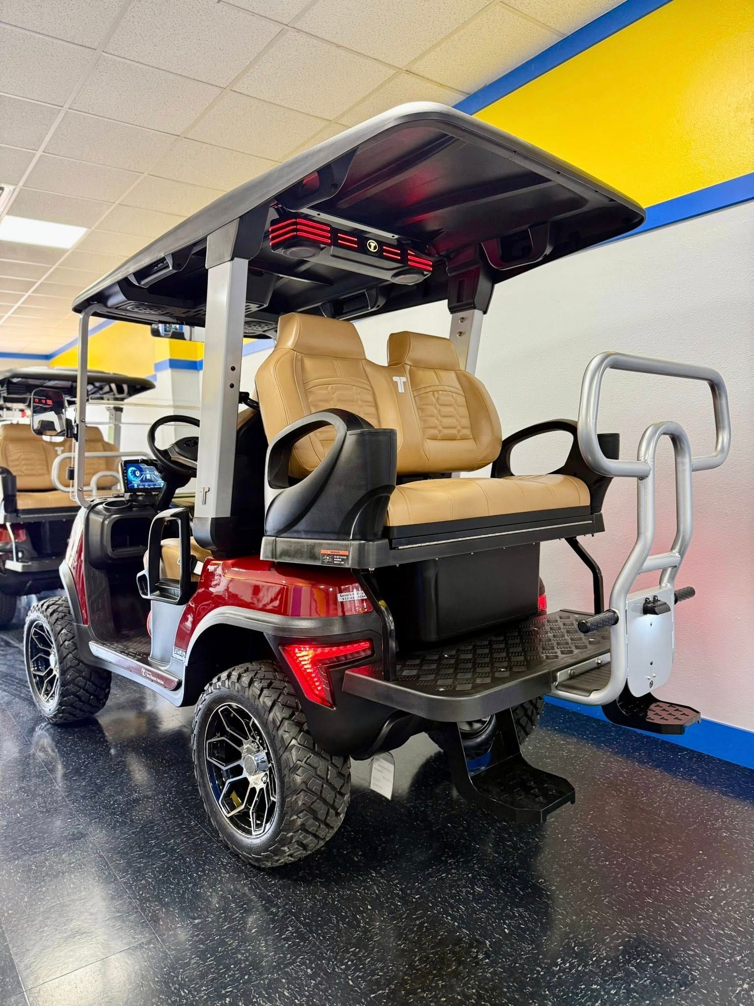 Red golf cart with tan seats and black roof, parked indoors. It has large black tires and a rear-facing seat.