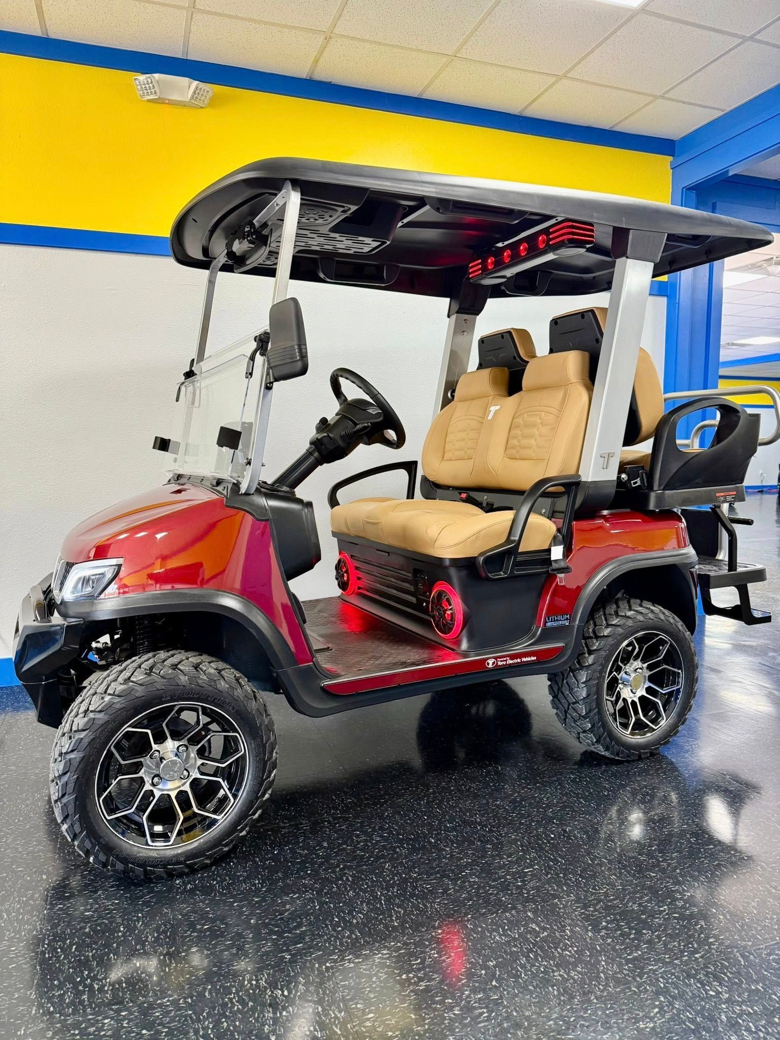 Red and black golf cart with tan seats and large black wheels. It is parked inside a building with yellow and blue accents.
