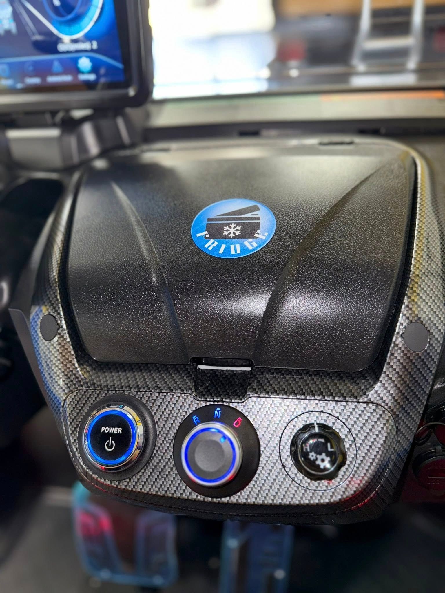 Dashboard with buttons and a screen. A black, textured storage compartment has a blue logo and is surrounded by a carbon fiber pattern.