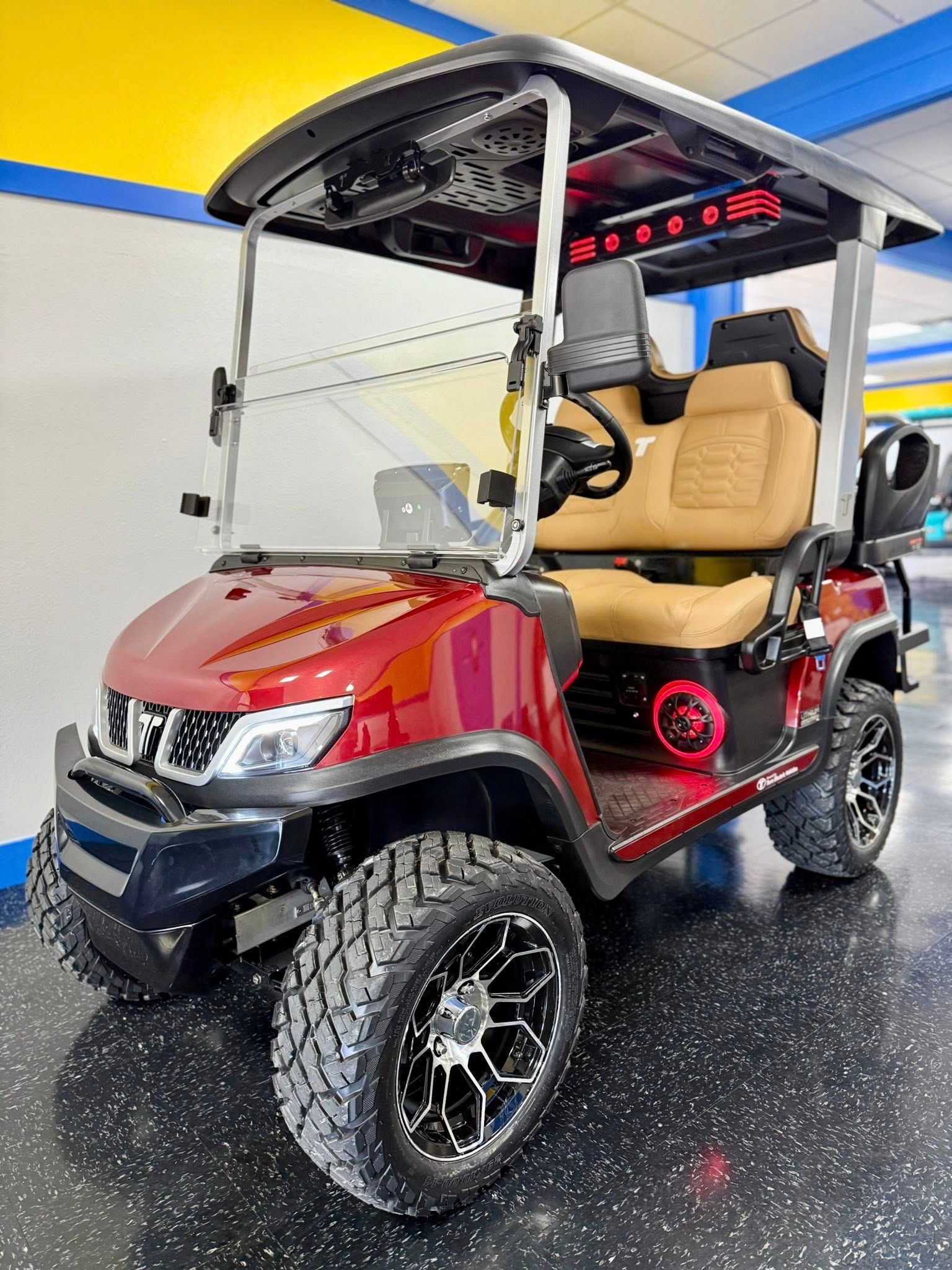 Red and black custom golf cart with off-road tires, tan seats, and a black roof, displayed inside a showroom.