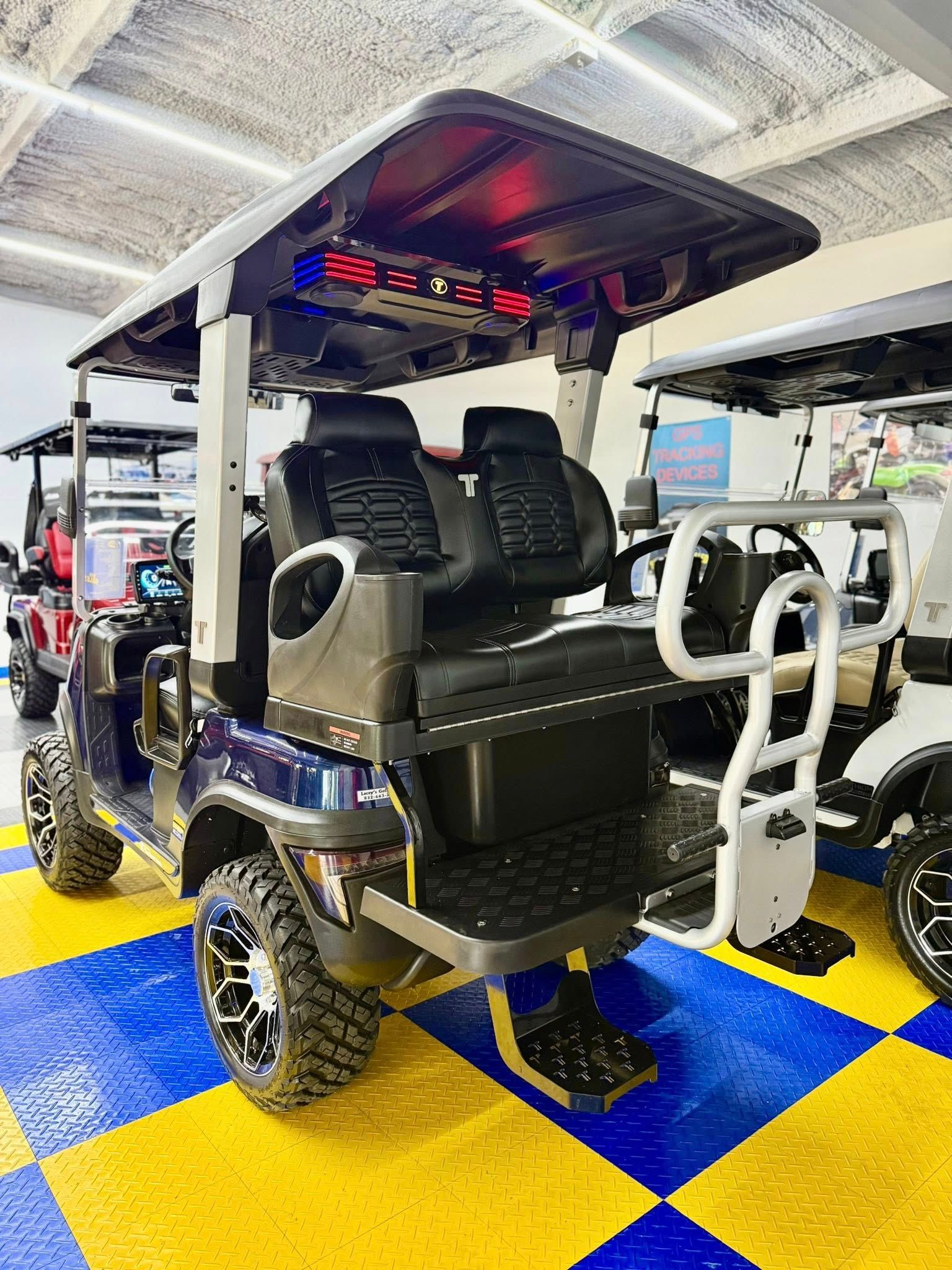 A dark blue golf cart with black seats and large wheels. It has a rear step, a roof with speakers, and is in a showroom.