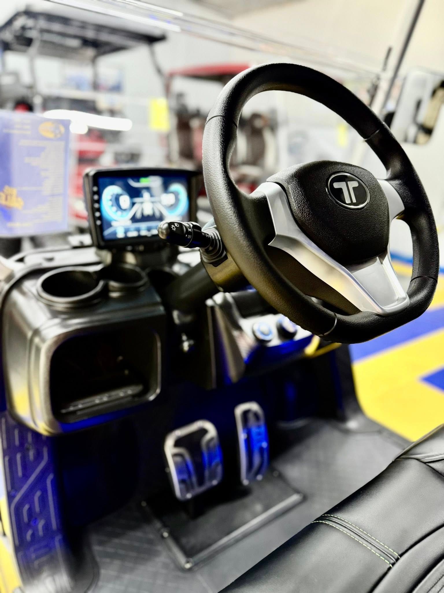 Interior of a golf cart: black steering wheel, digital display, and chrome accents. Dark upholstery and blue and yellow flooring are visible.