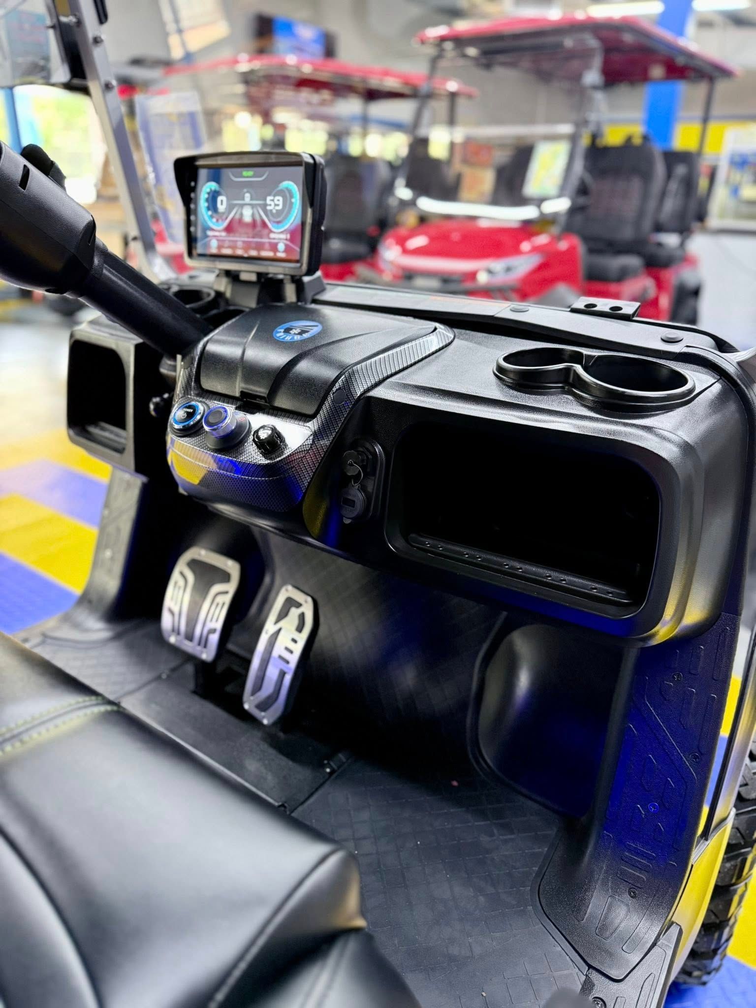Black golf cart interior with digital display, cupholders, and silver pedals. Two red golf carts are in the background.
