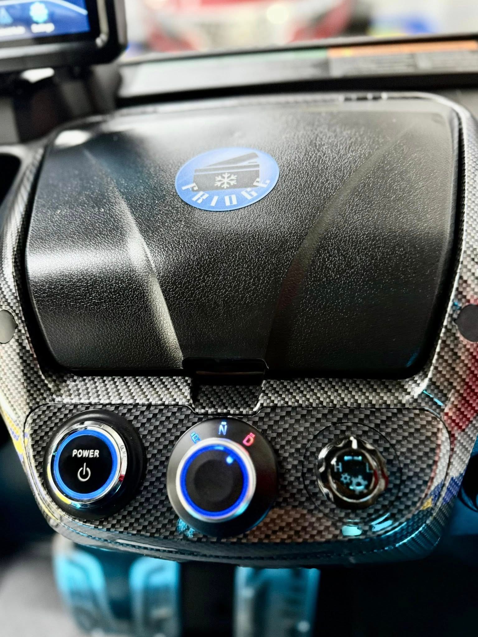 Dashboard close-up with carbon fiber texture, three illuminated controls, and a blue emblem.