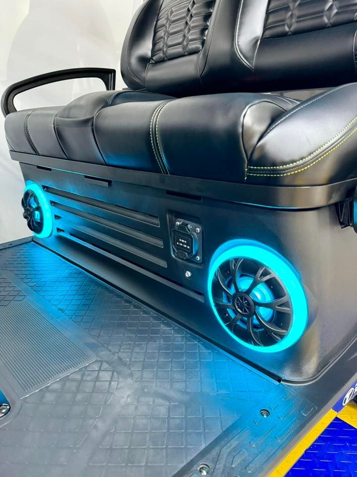 Black golf cart seat with blue LED-lit speakers and yellow stitching. Gray floor with blue and yellow accent.