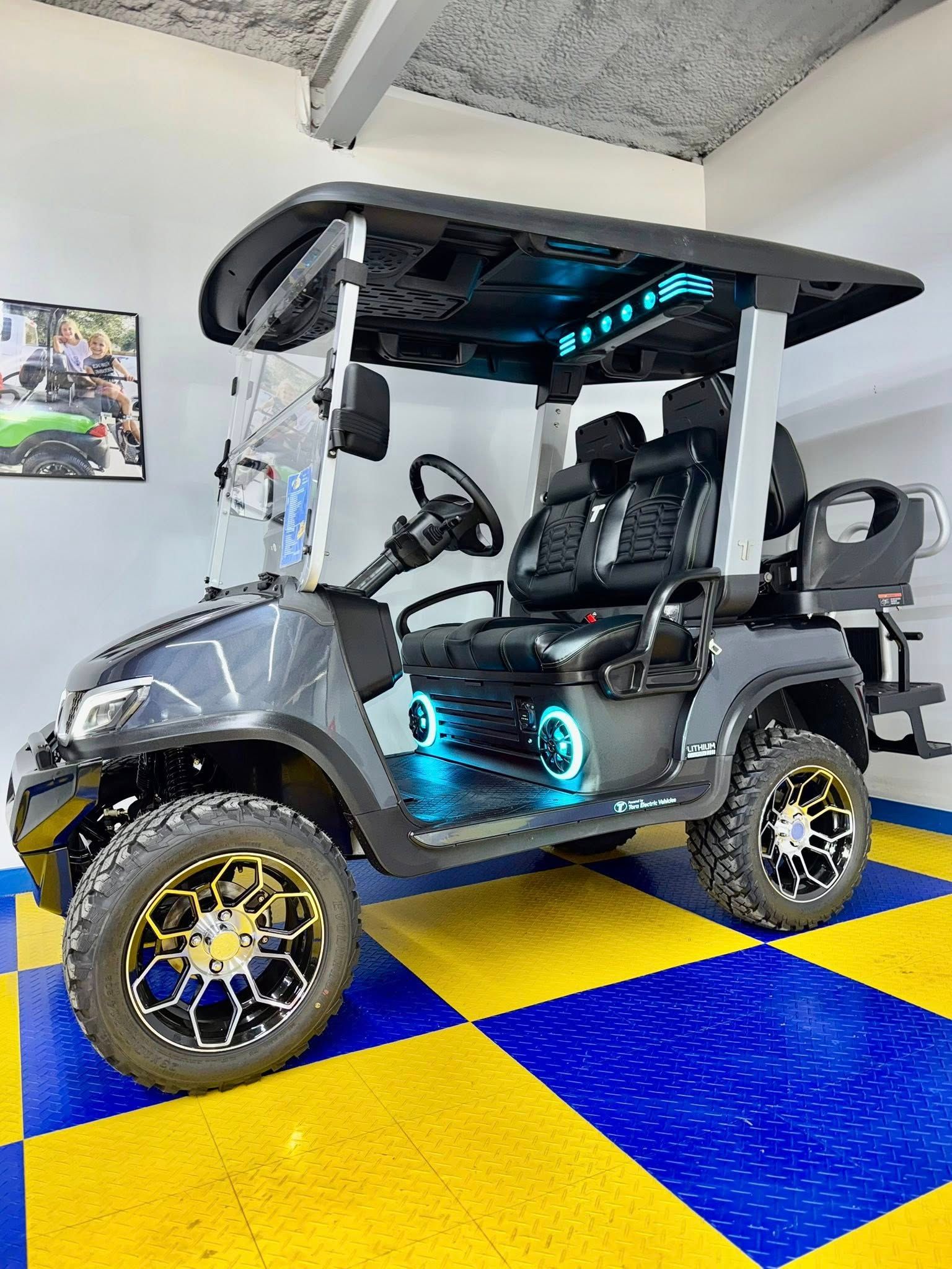 Customized gray golf cart with blue LED lighting, off-road tires, and a black roof, parked on a yellow and blue checkered floor.