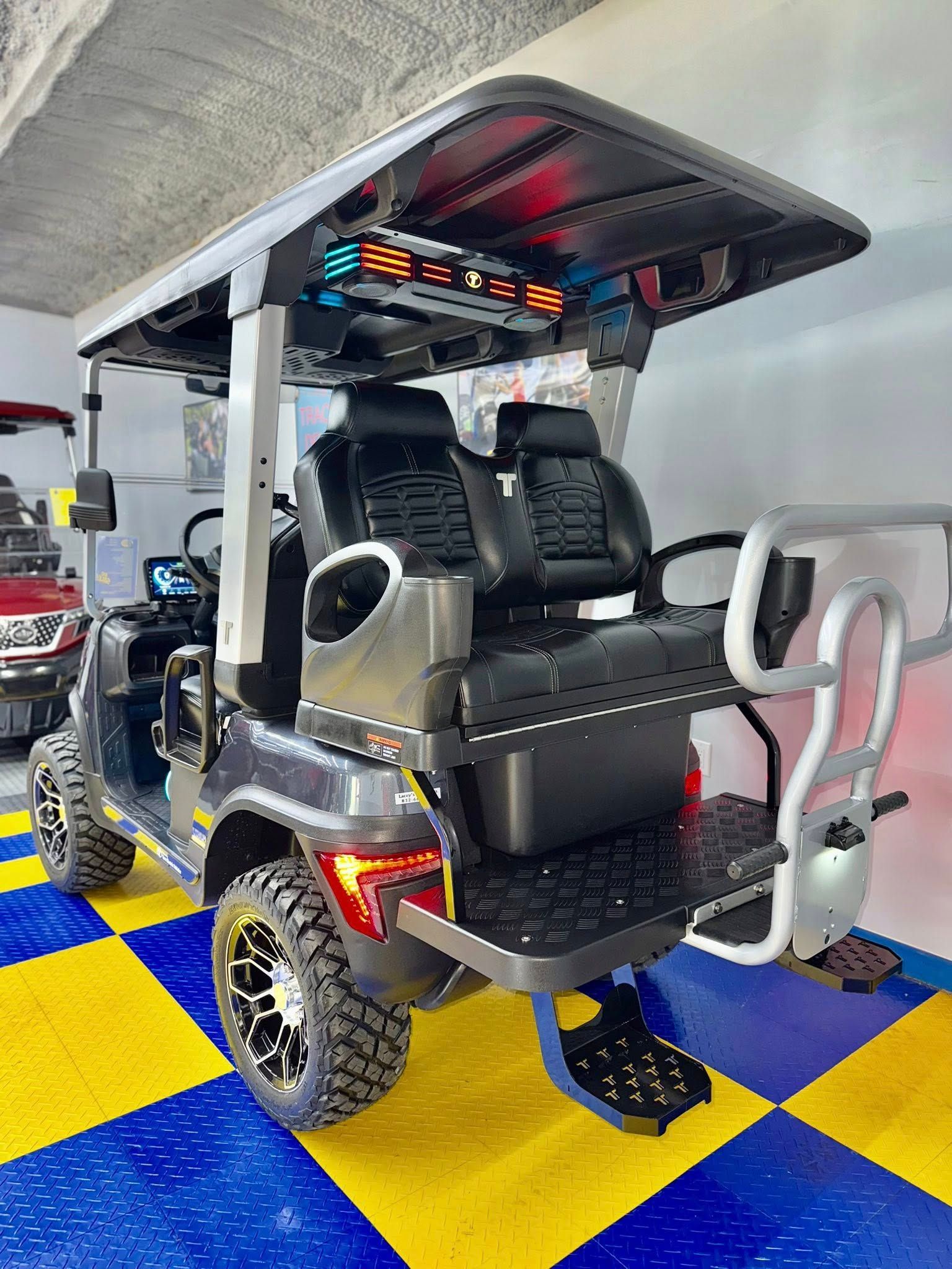Dark gray golf cart with black seats, rugged tires, and rear-facing bench seat. It's parked on a yellow and blue checkered floor.