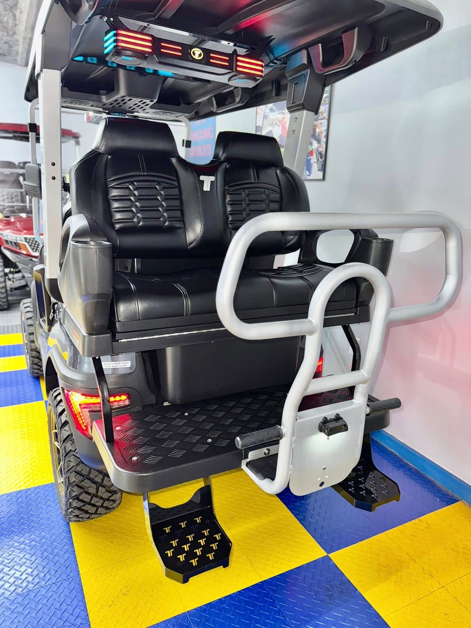 Back view of a black golf cart with a rear-facing seat, mounted on a blue and yellow checkered floor.