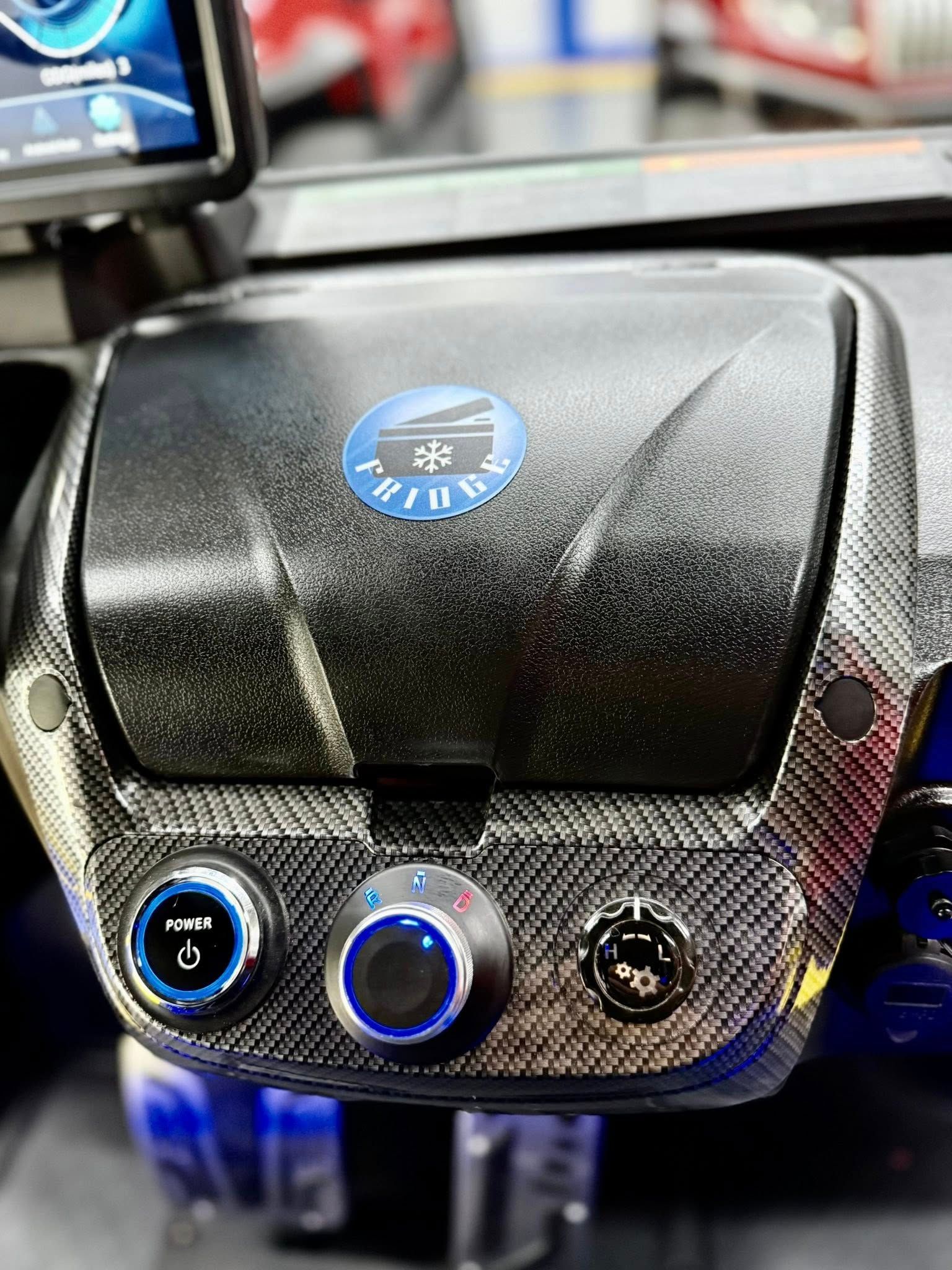 Close-up of a car dashboard with carbon fiber texture, three control knobs with blue lights, and a logo that reads