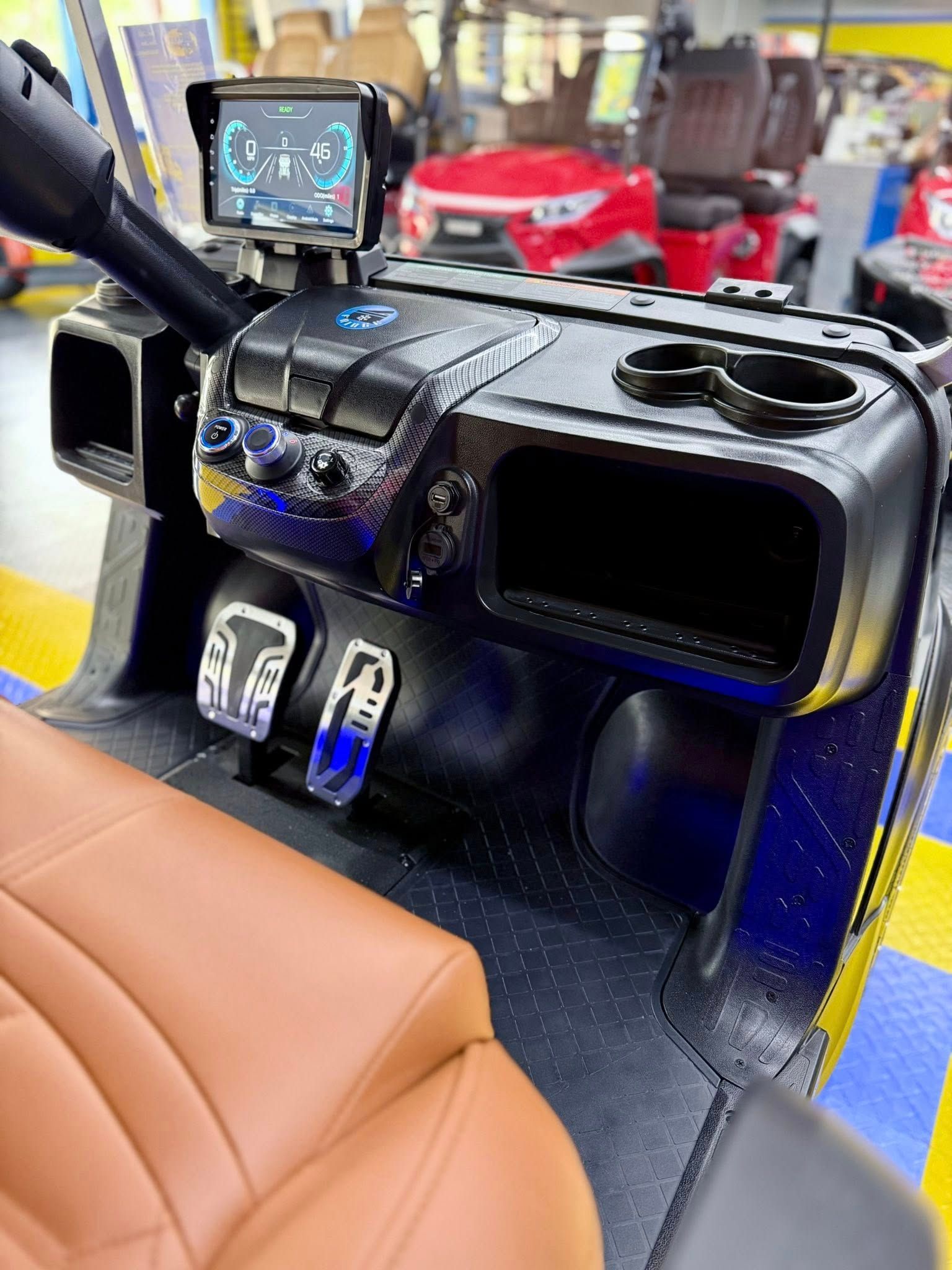 Interior of a golf cart featuring a black dashboard with an LCD screen, cup holders, pedals, and brown leather seating.