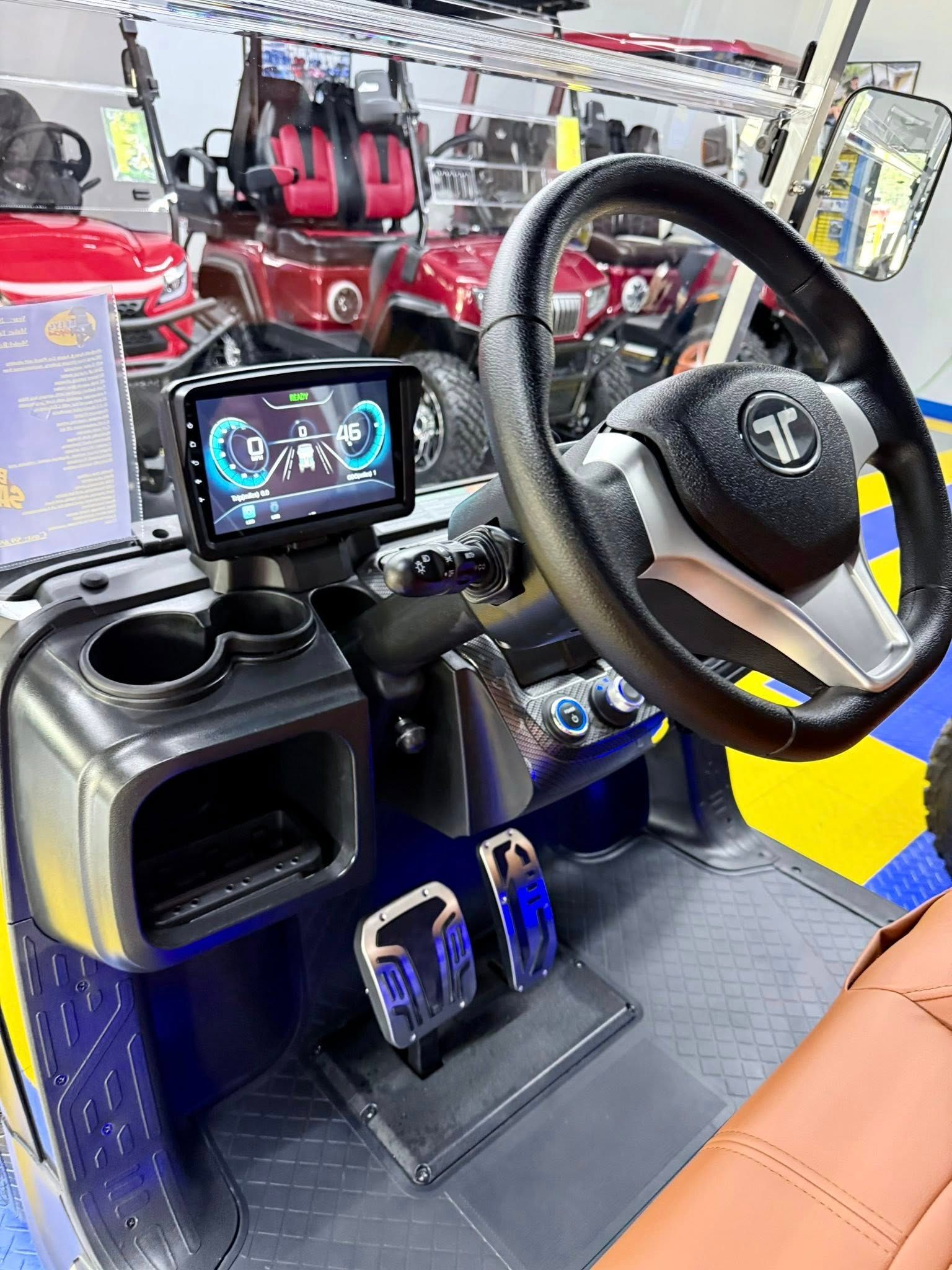 Interior of a golf cart featuring a black steering wheel, digital display, blue and silver trim, and brown seating.