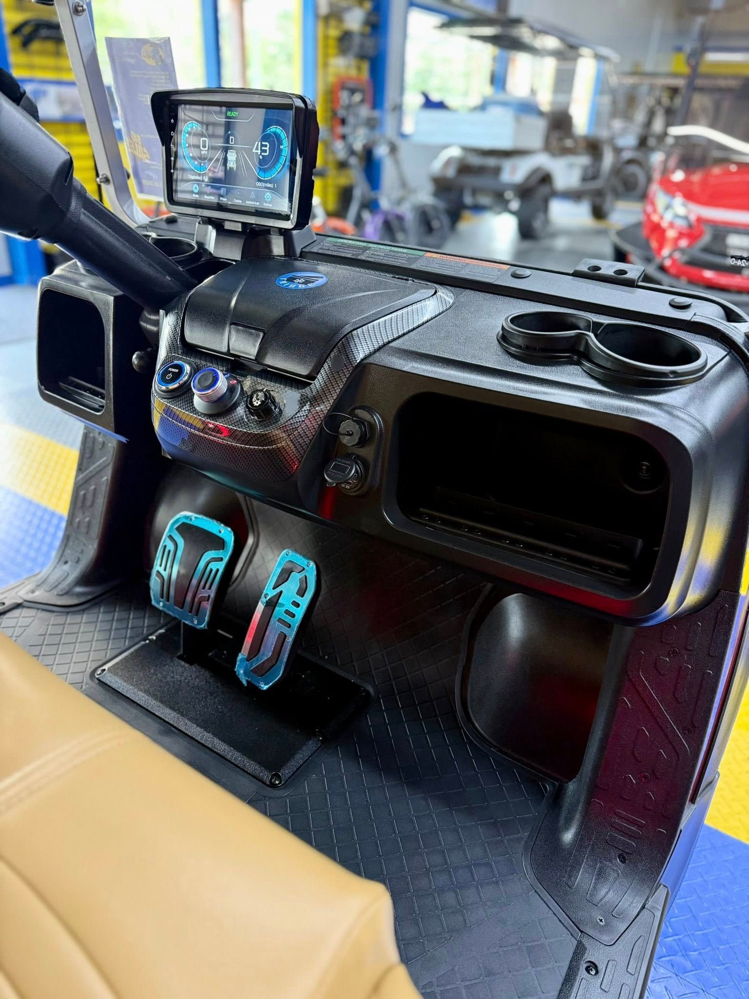 Close-up of a golf cart's black dashboard, with a screen, cup holders, pedals, and tan seat visible.