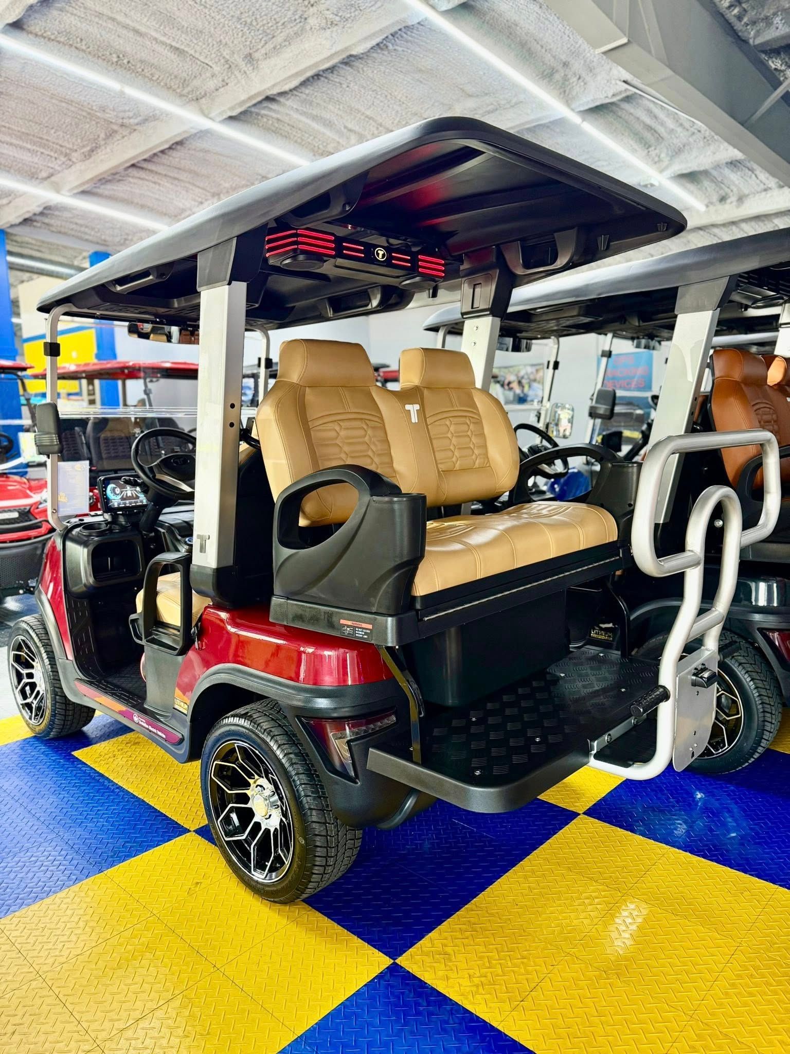 Red and tan golf cart with custom wheels and seats, parked on a checkered blue and yellow floor, likely a showroom.