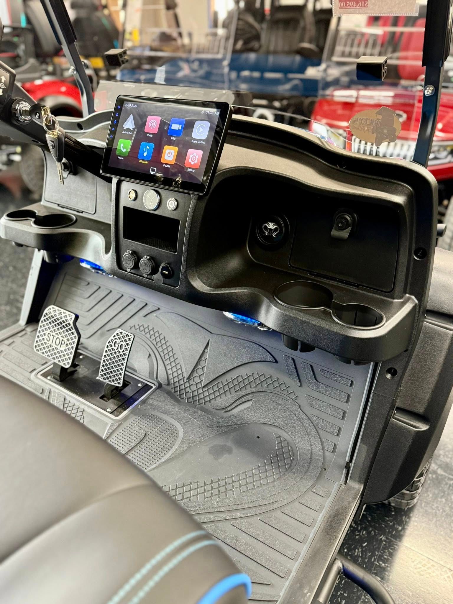 Golf cart interior: black dashboard with touchscreen, cupholders, pedals, and rubber mat with tire tread pattern.
