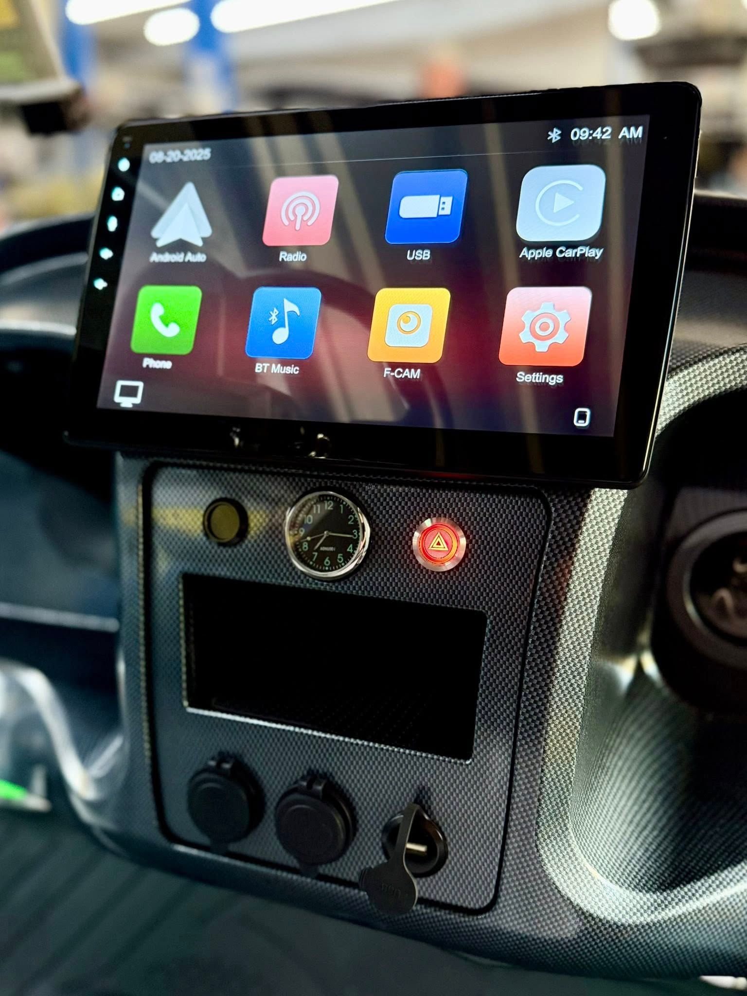 Car dashboard with touchscreen display, buttons, and power outlets. Dark gray panel with colorful app icons.