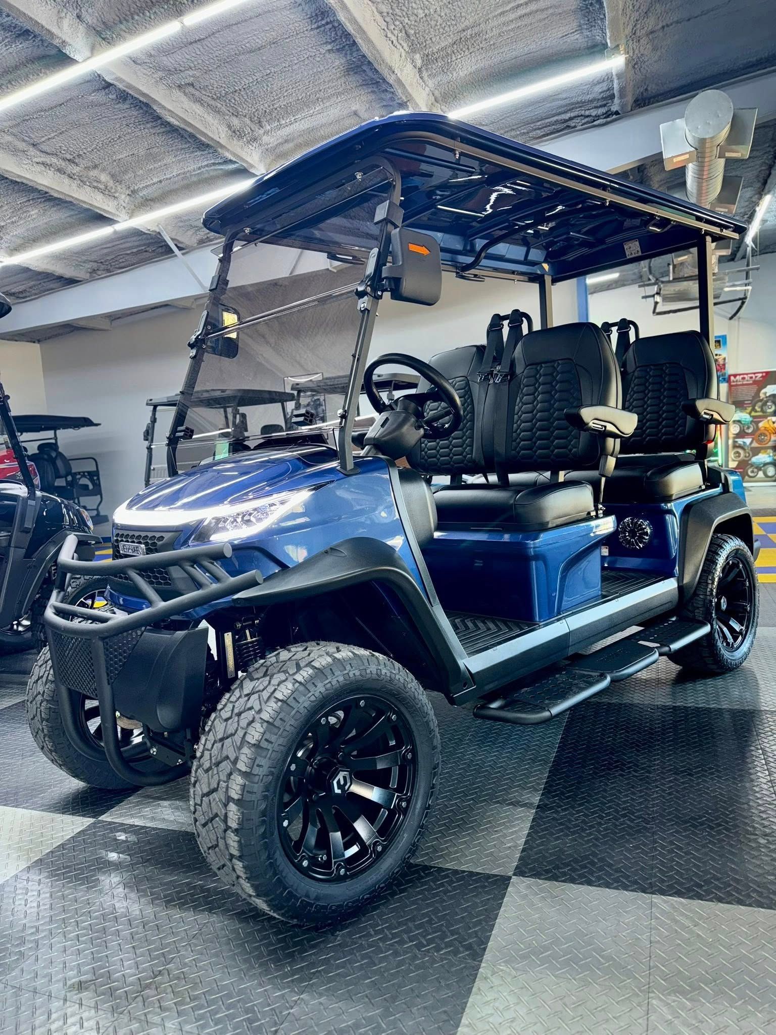 Blue golf cart with black wheels, roof, and accessories. Interior setting with checkered floor.