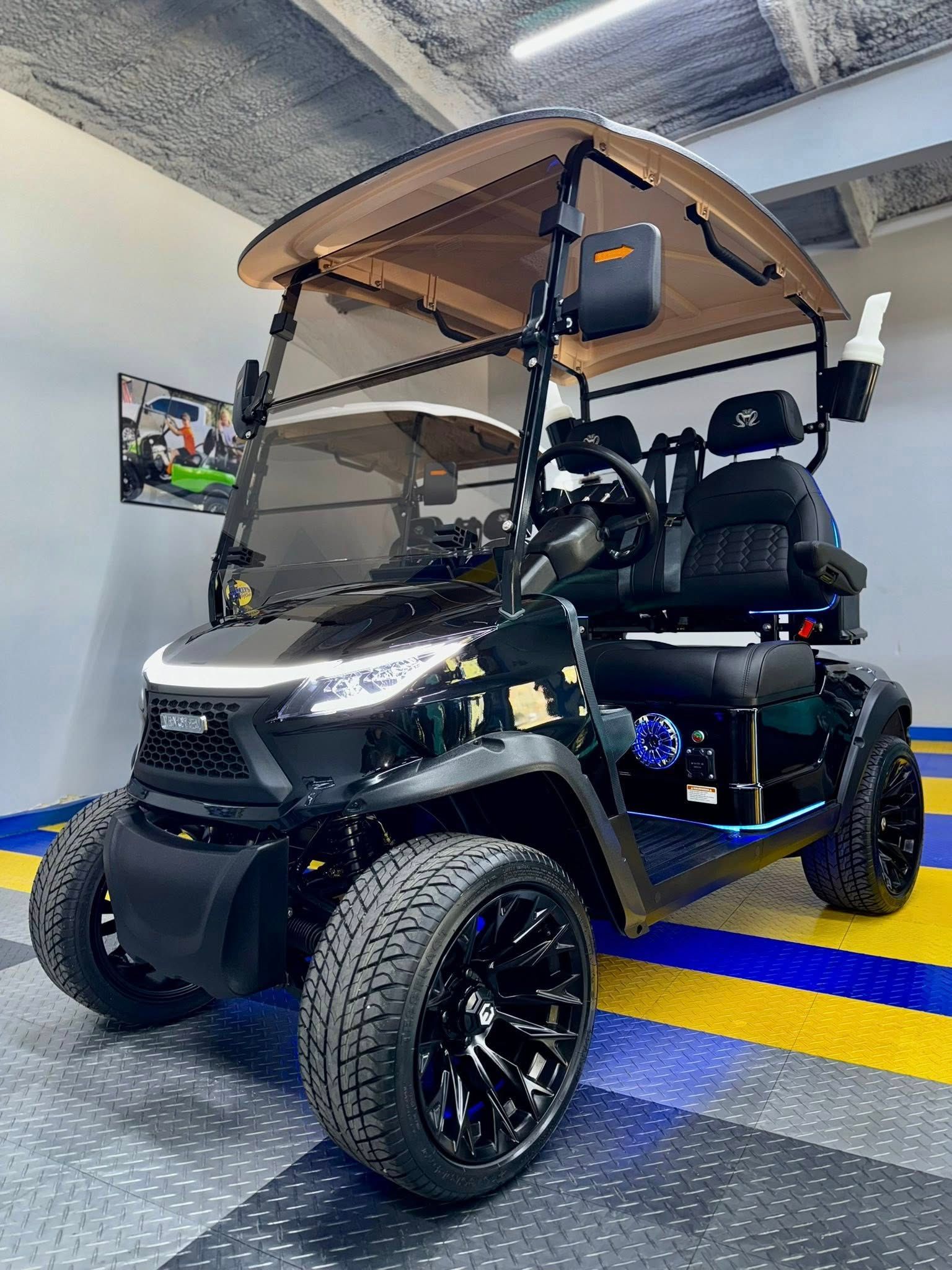 Black golf cart with black rims and large tires, tan roof, parked indoors.