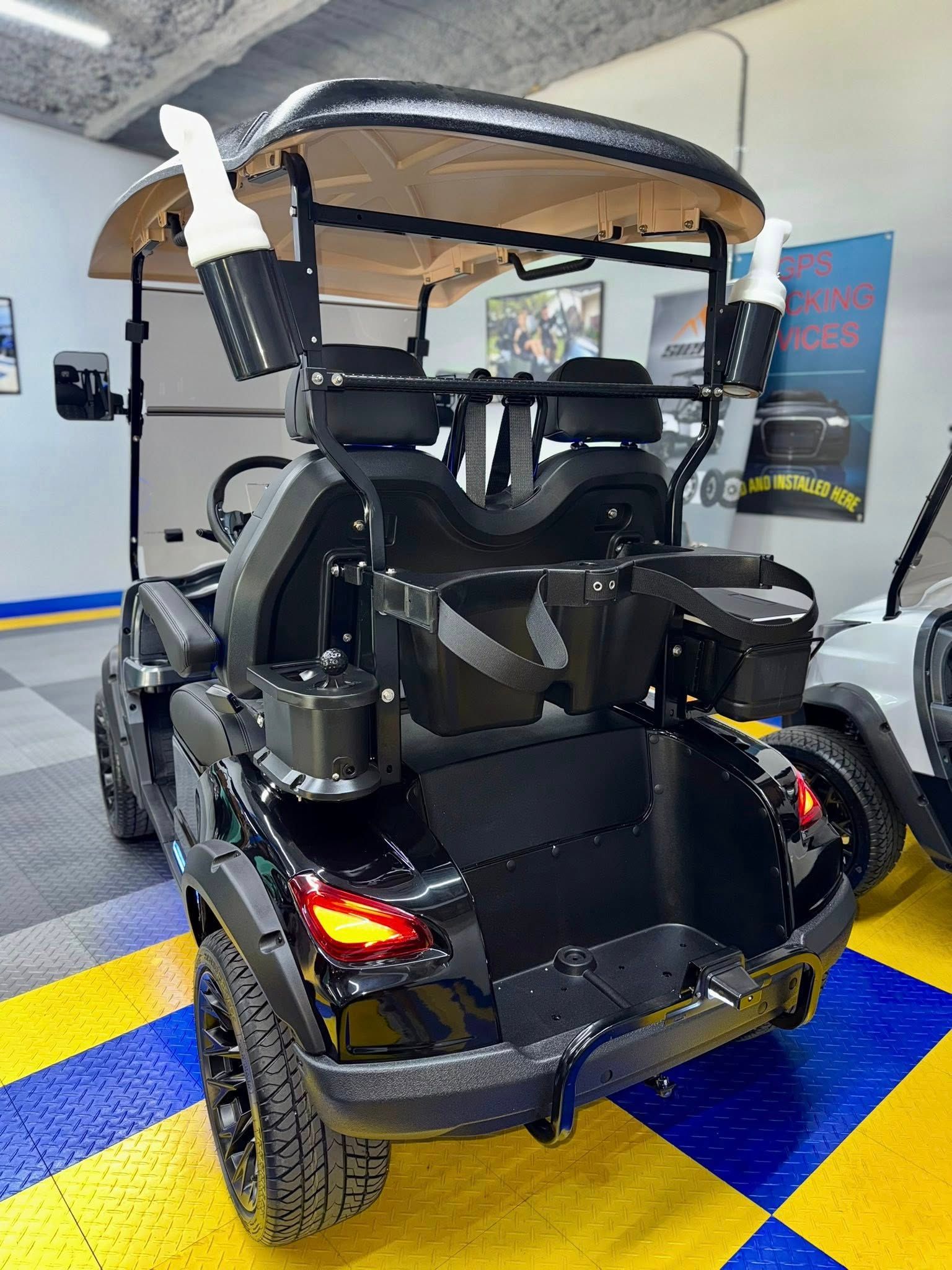 Black golf cart with off-road tires, cup holders, and a rear bench. Indoors, tile floor.