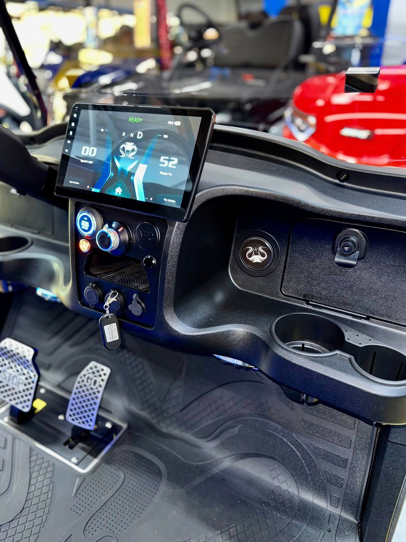 Dashboard of a golf cart with a screen displaying speed, black, textured interior, cup holders, and foot pedals.