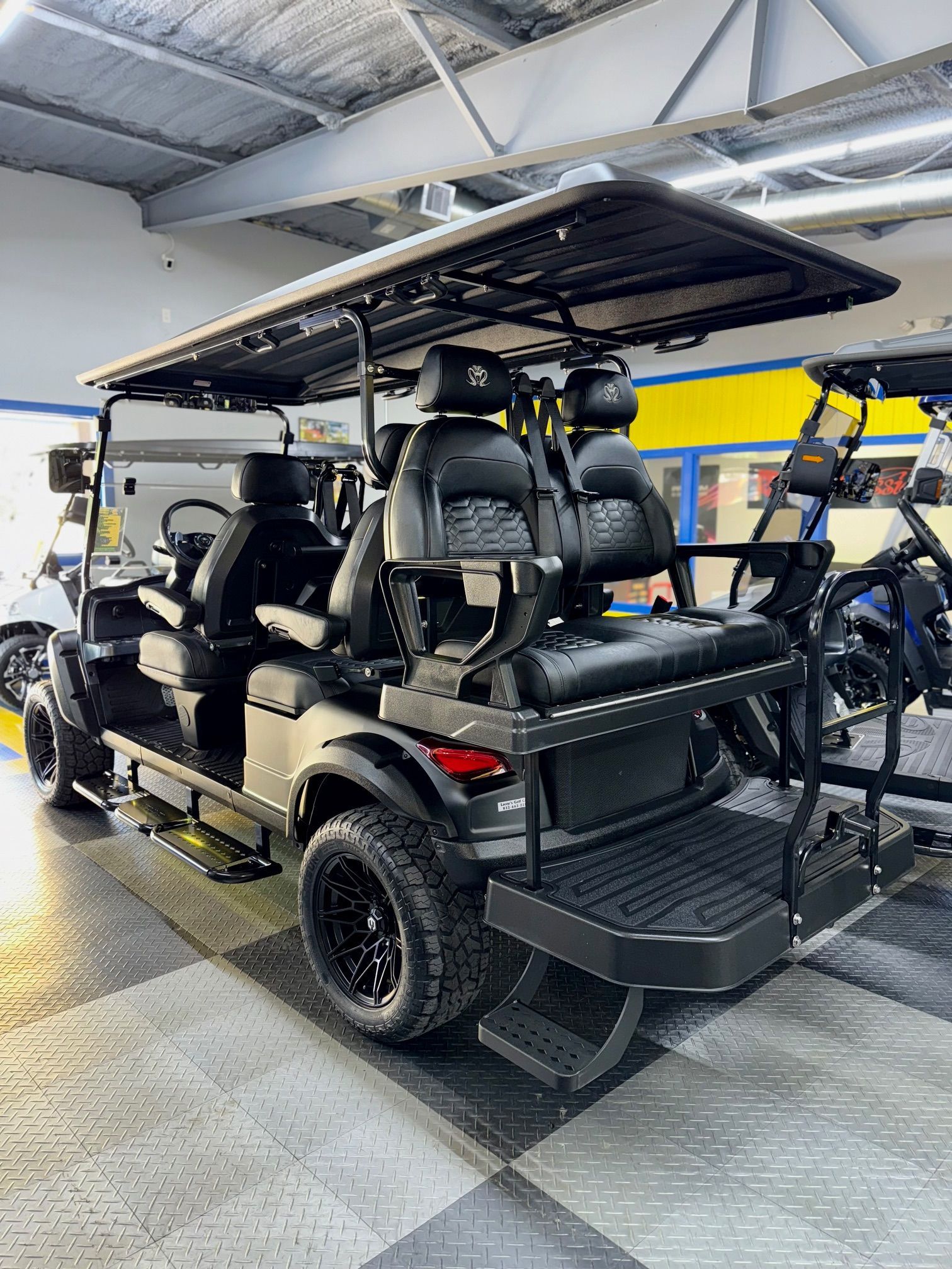 Black six-passenger golf cart with black rims and roof rack, parked indoors on a tiled floor.