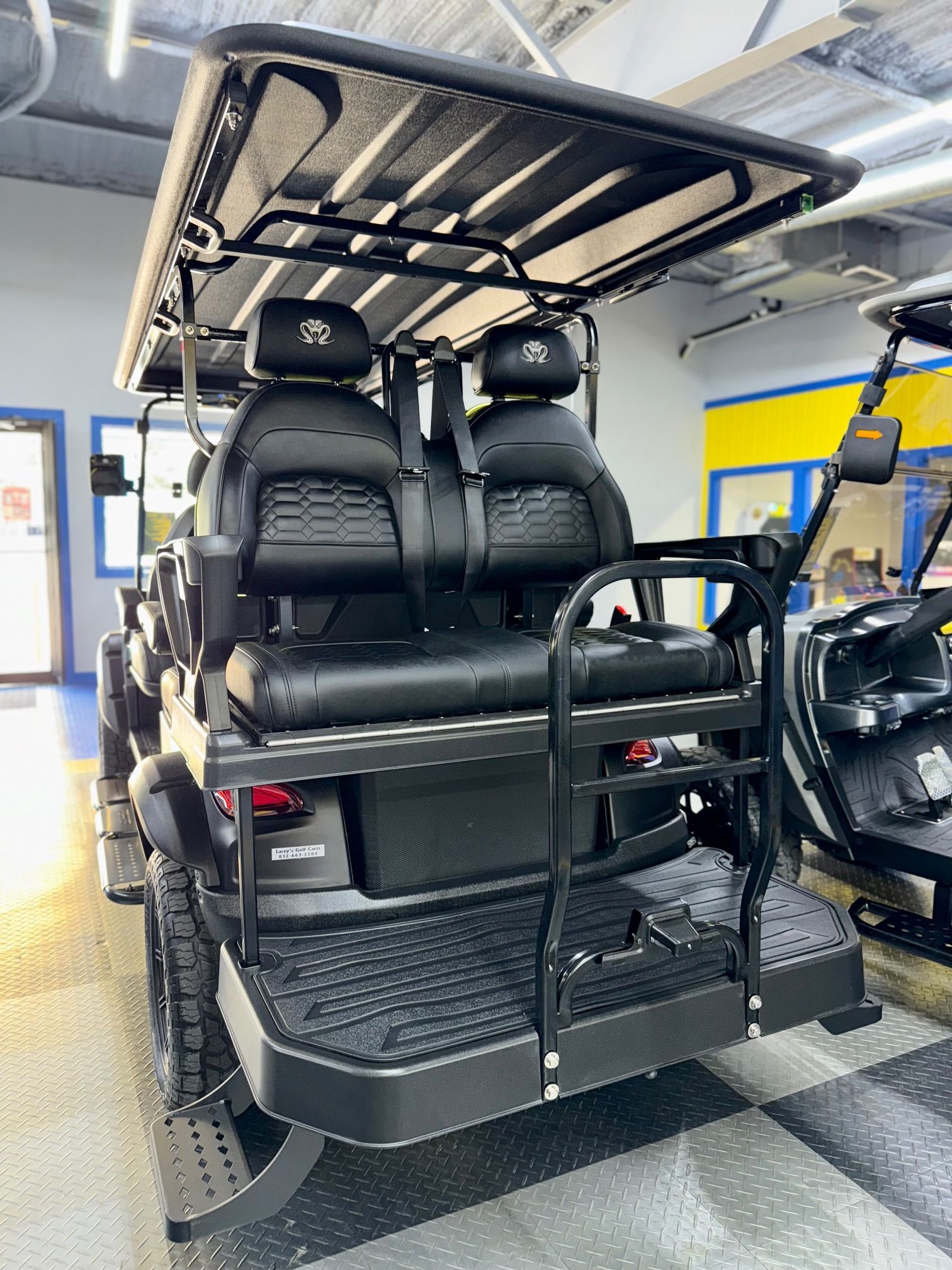 Black golf cart with back seating, roll bars, and black tires, inside a building.