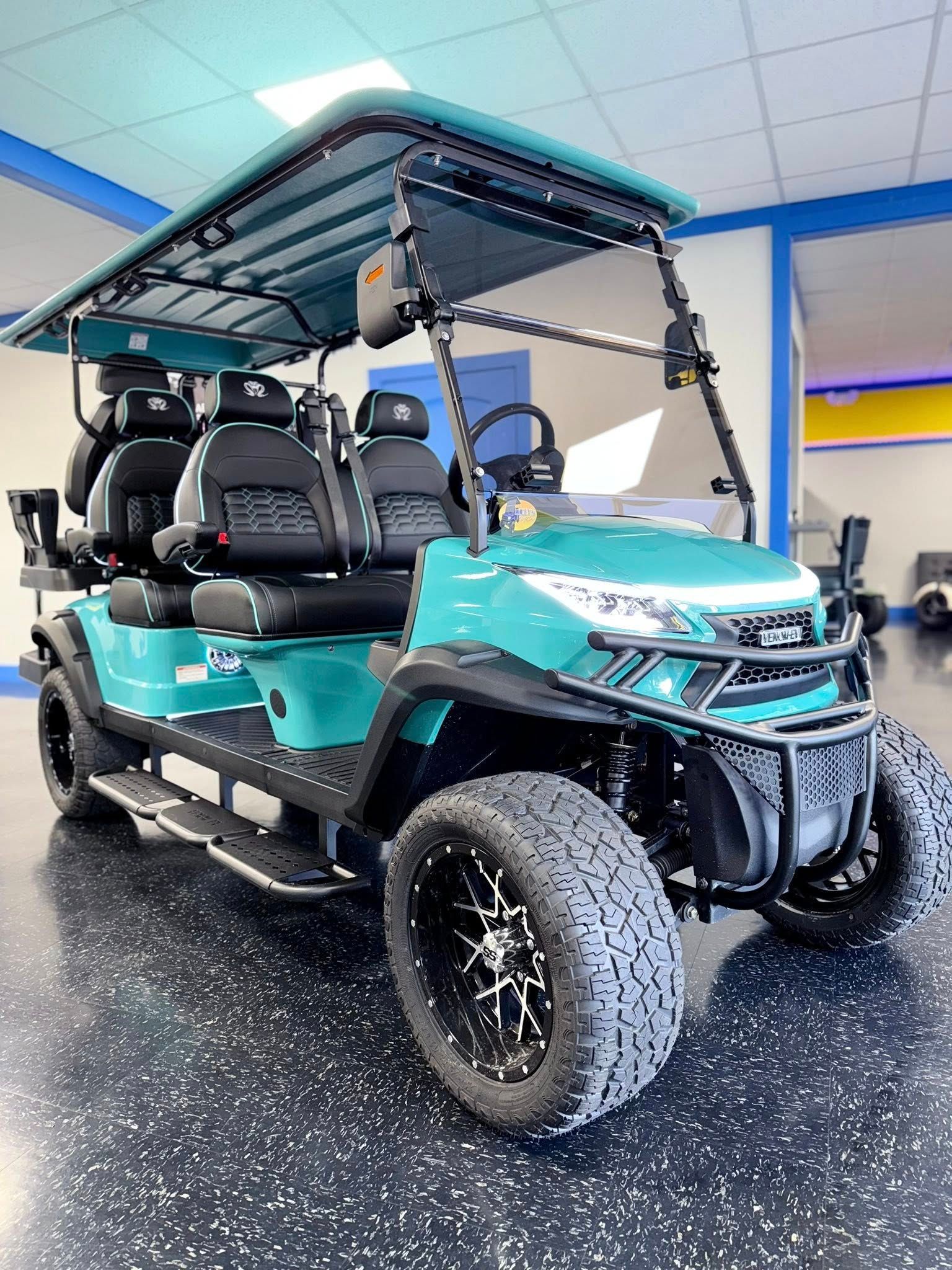 Teal 6-passenger golf cart with black accents, black wheels, and off-road tires. Indoor setting.