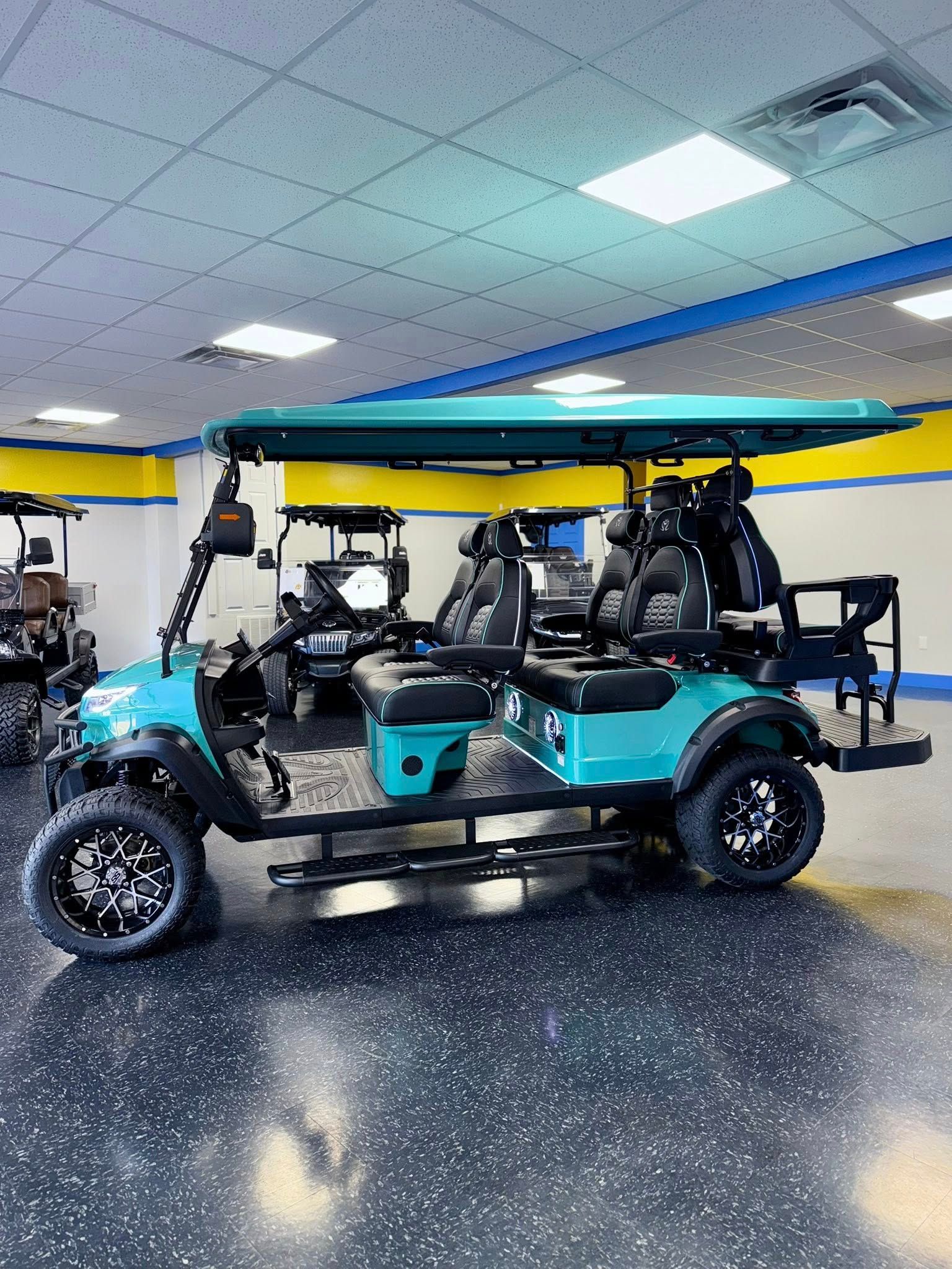 Teal six-seat golf cart with black accents, large tires, and a teal roof, parked indoors.