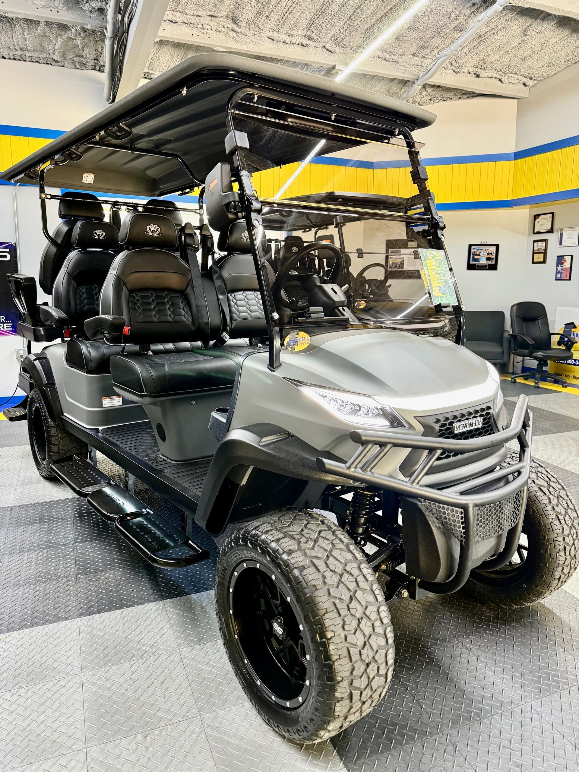 Gray golf cart with black accents, rugged tires, and a roof rack, inside a showroom.