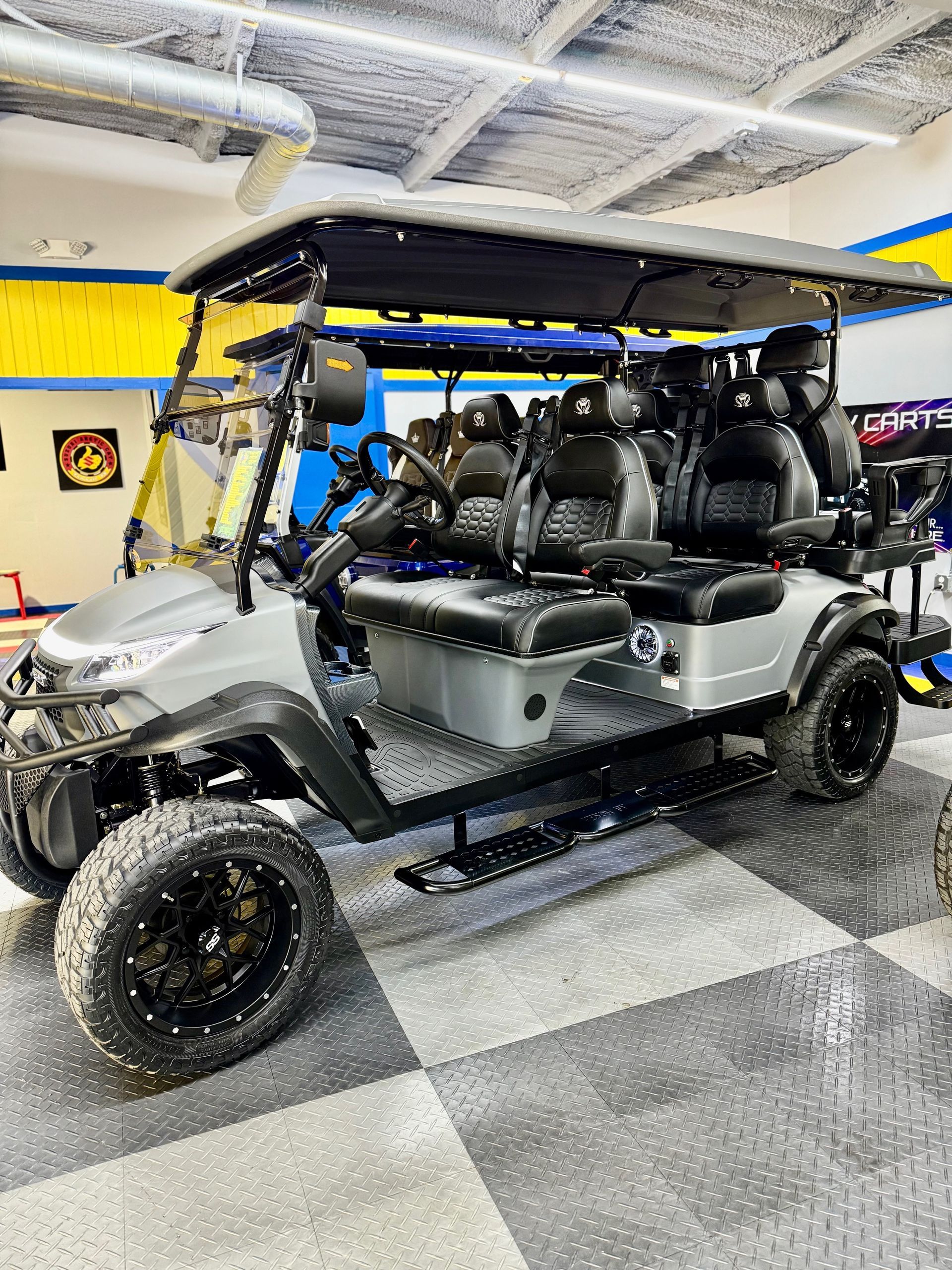 Gray lifted golf cart with black wheels, seats, and roof, parked on checkered floor.