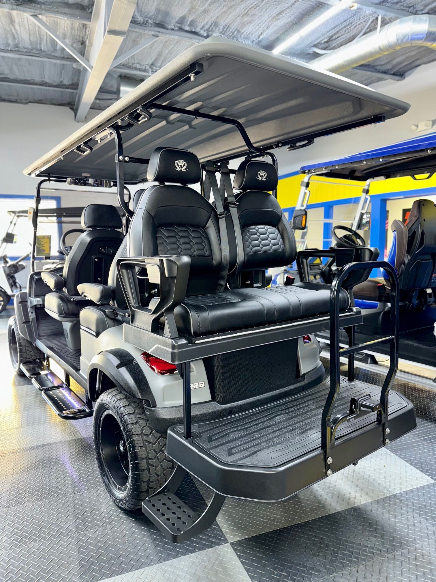 Gray golf cart with black seating, lifted wheels, and rear step. Inside a building.