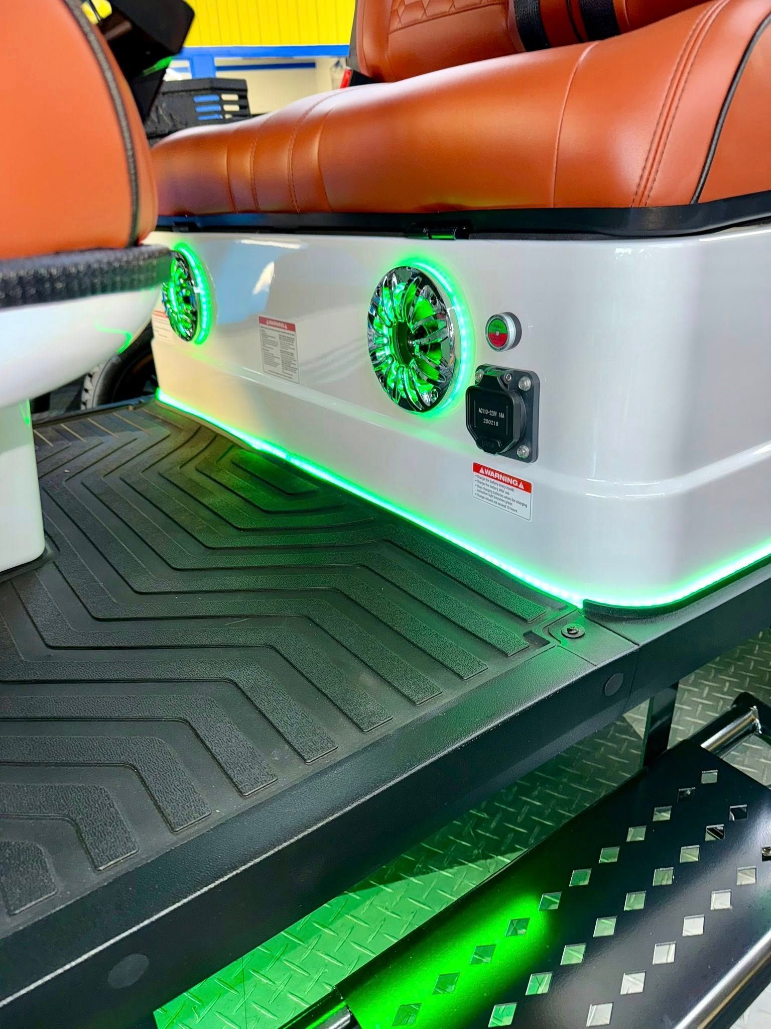 Close-up of a white cart audio box with green LED lighting, speakers, and a power outlet, next to brown seating.