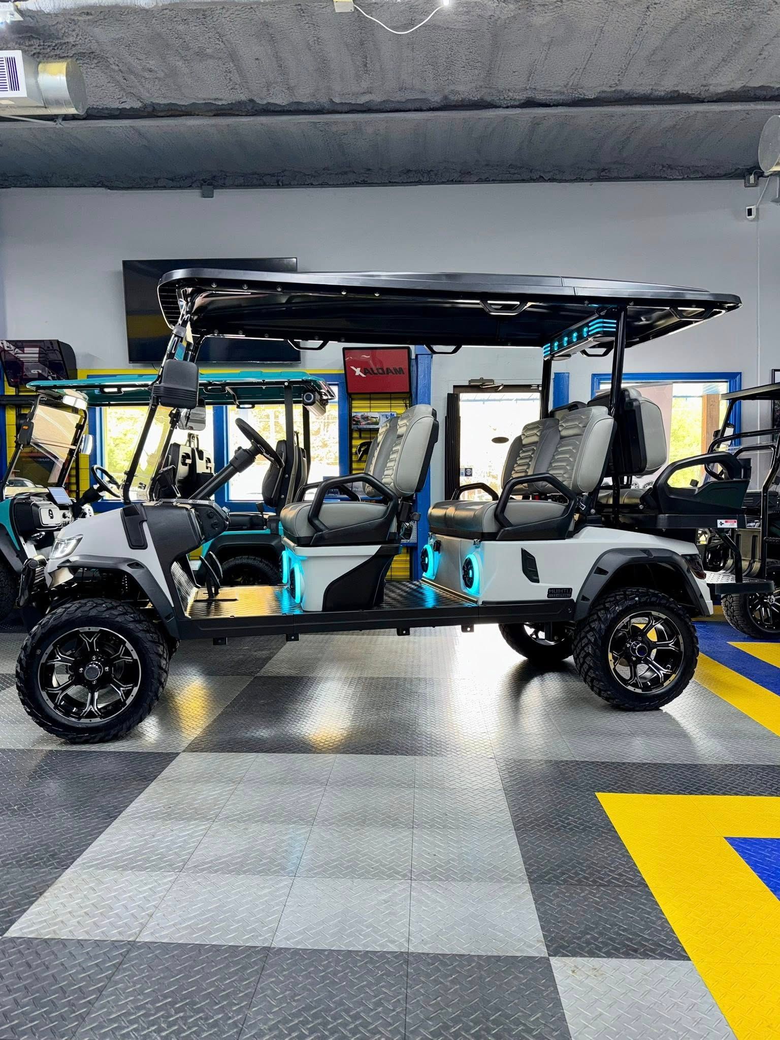 Gray and black six-seater golf cart with off-road tires, parked indoors on checkered flooring.