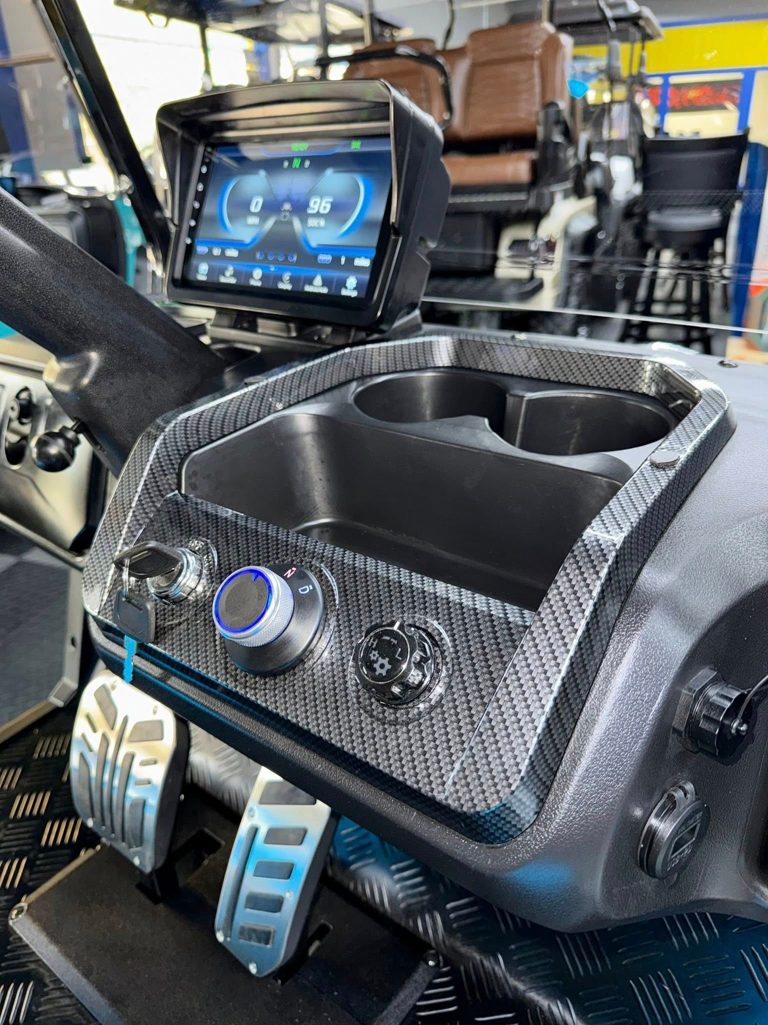 Golf cart interior: carbon fiber console with cupholders, digital display, pedals, and blue accent lighting.