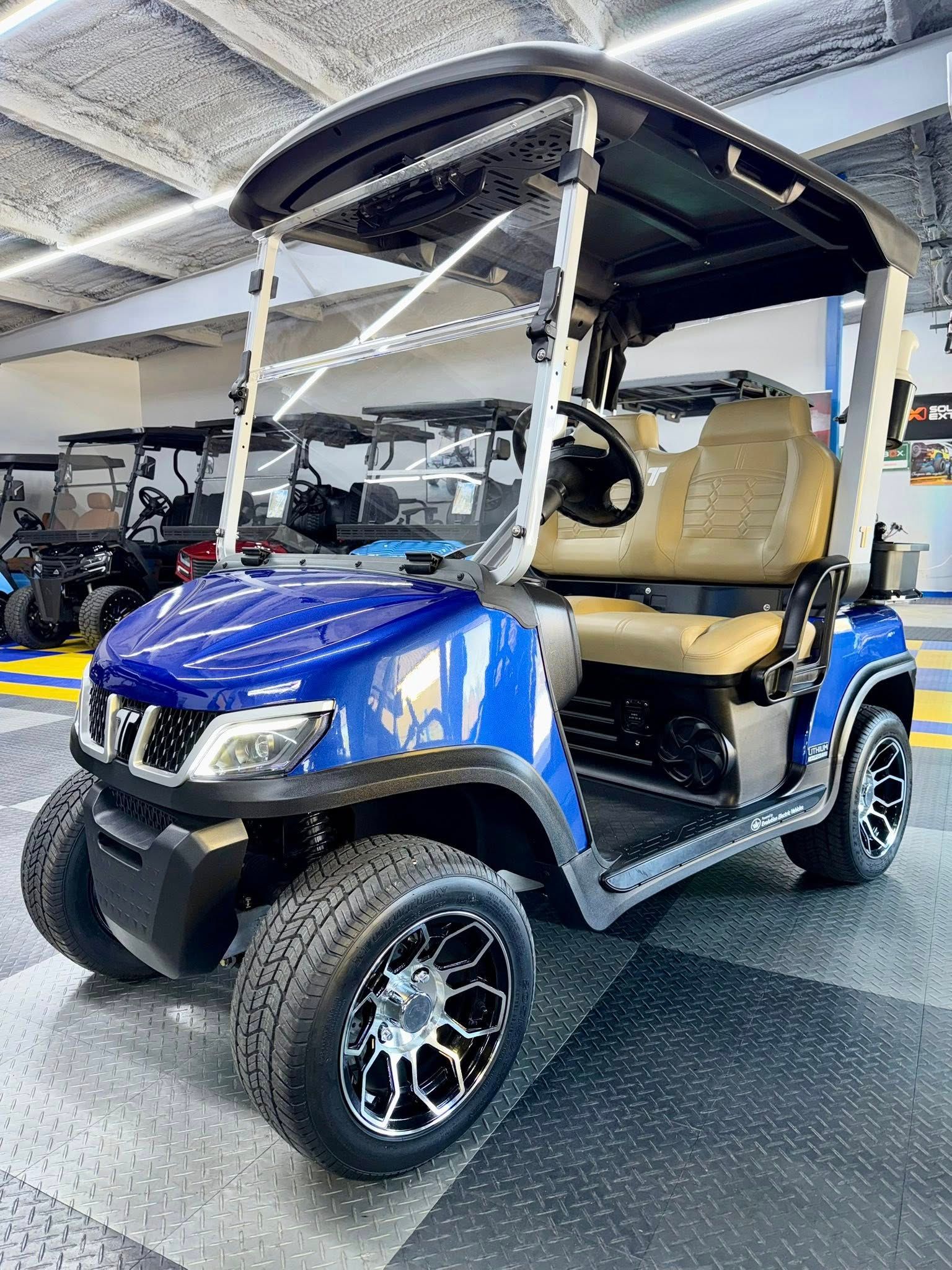 Blue golf cart with tan seats, black wheels, under canopy, indoors.