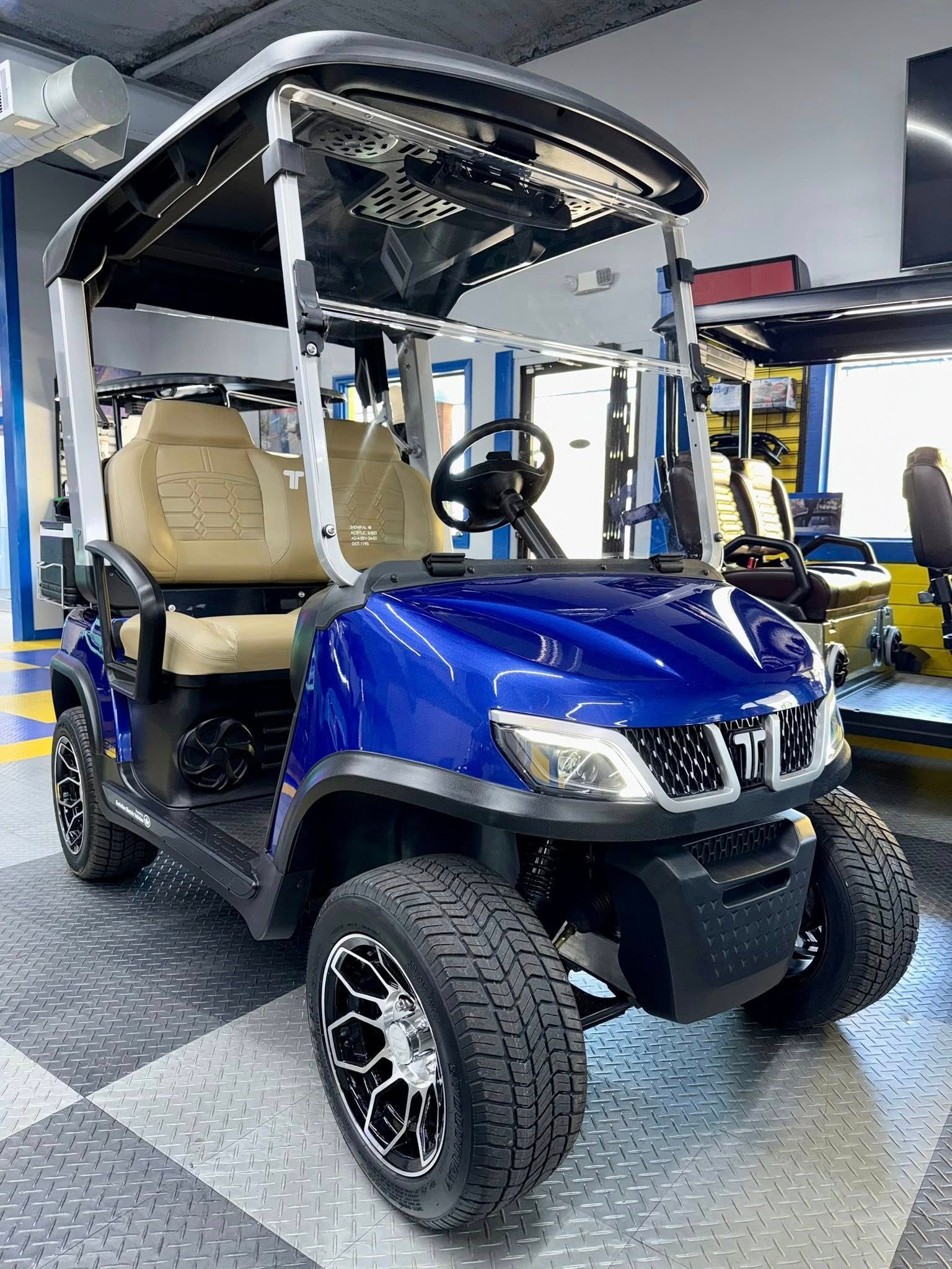 Blue golf cart with black roof, lifted tires, and chrome wheels on display indoors.