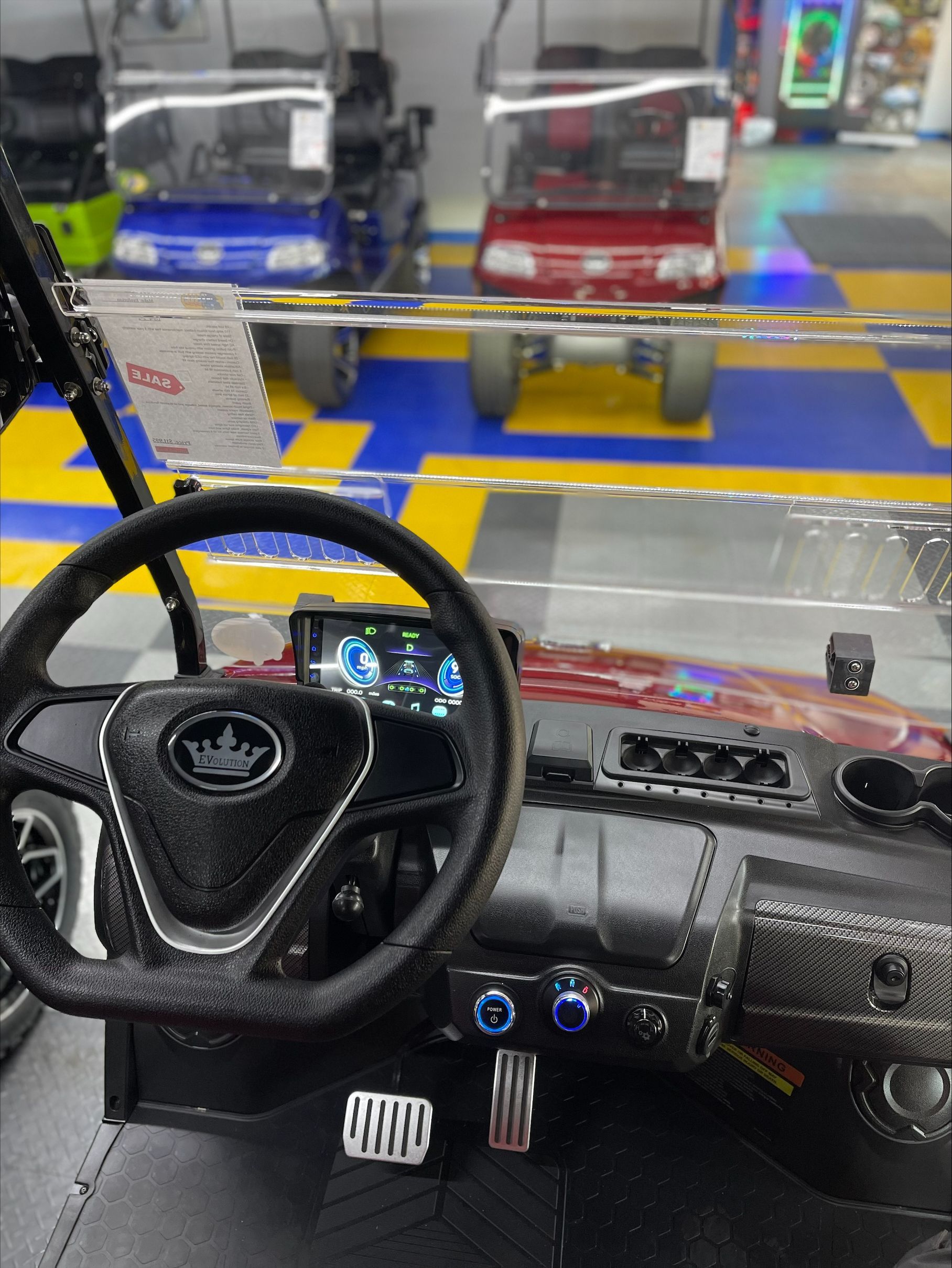 Interior view of a red golf cart, showing the steering wheel and dashboard. Other carts in blue, red, and green are visible in the background.