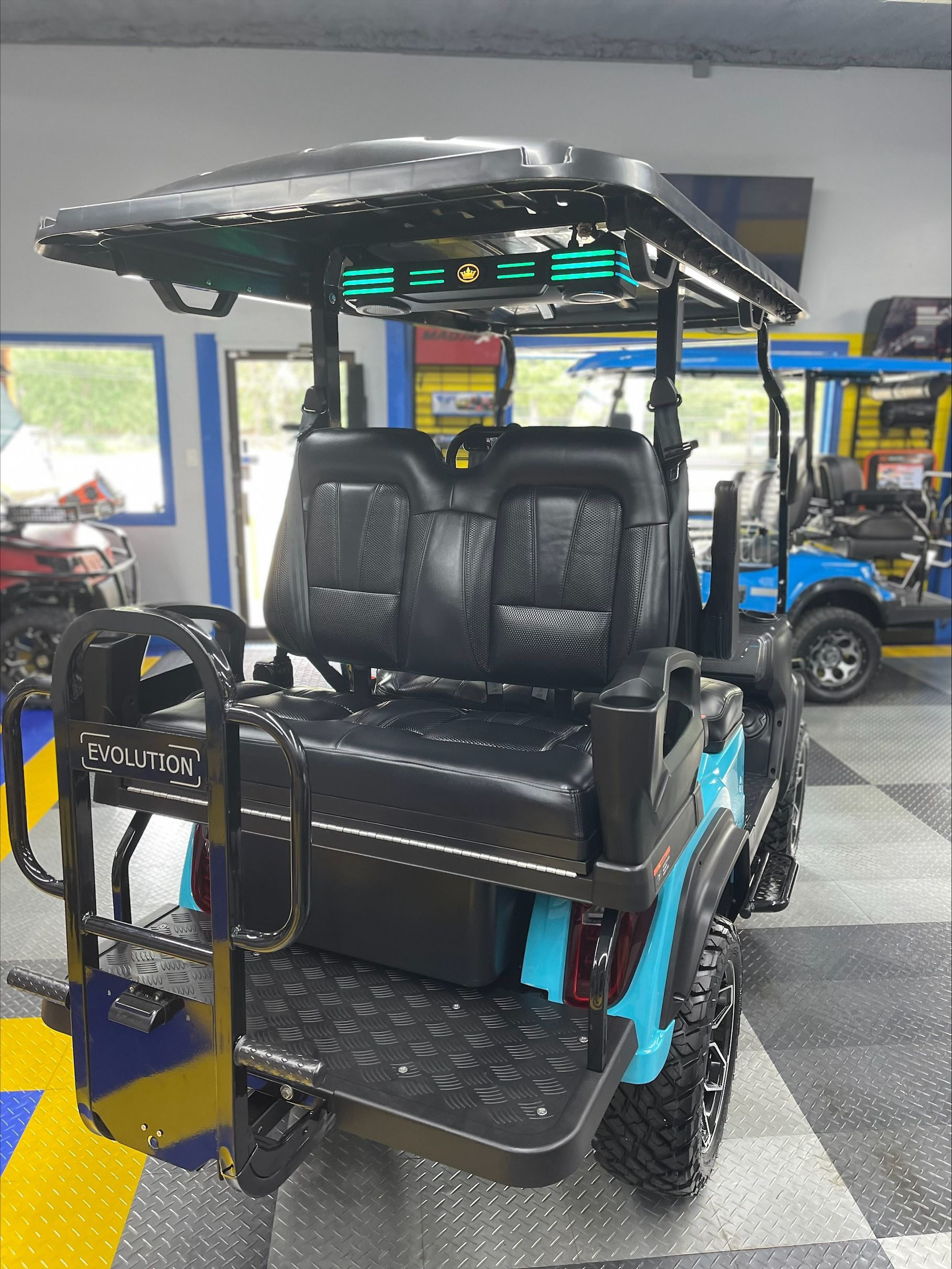 Blue golf cart with black seats and roof, parked indoors. The cart has large wheels and a rear-facing seat.