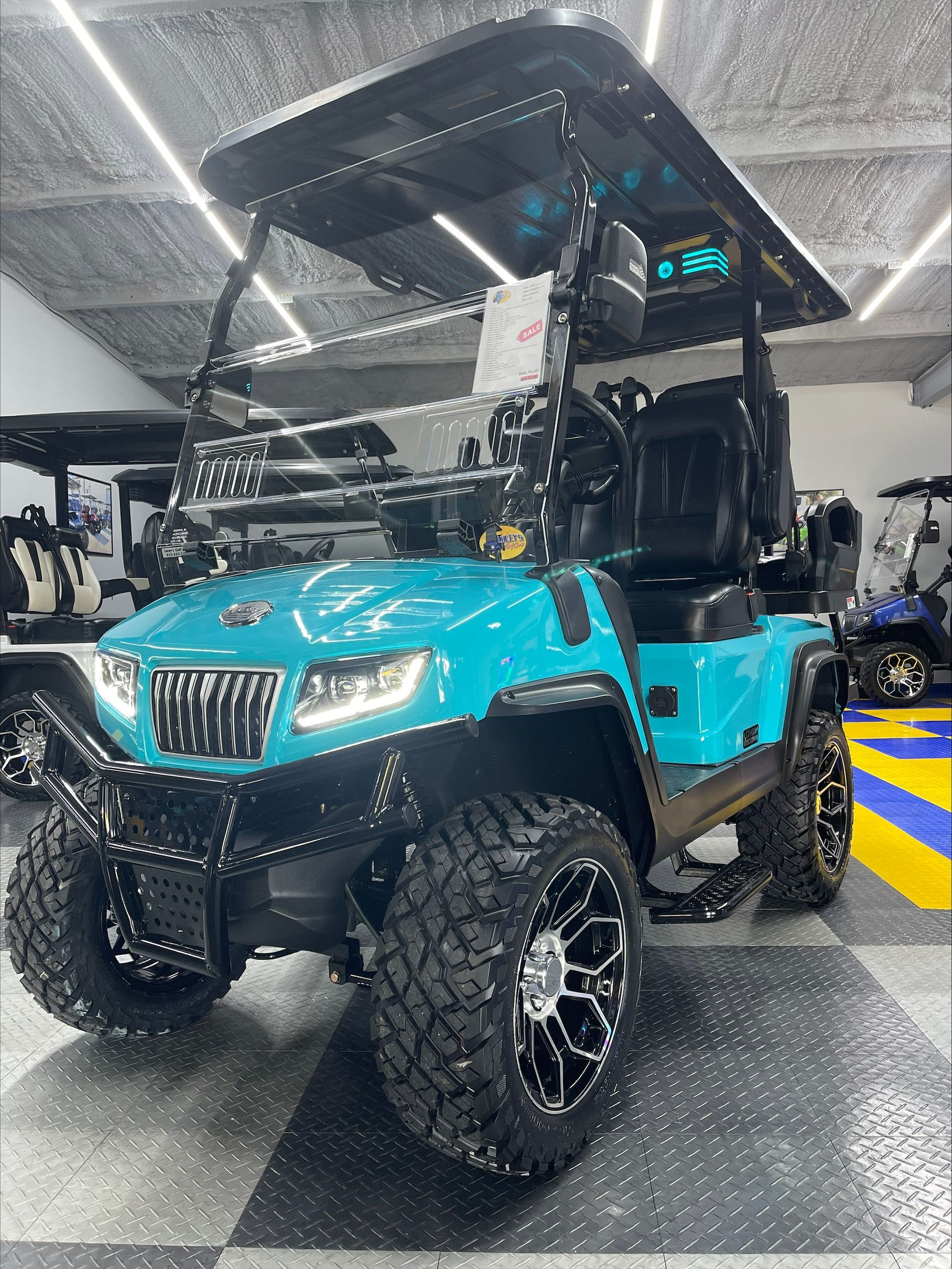 A turquoise golf cart with a black roof and large off-road tires is displayed in a showroom.