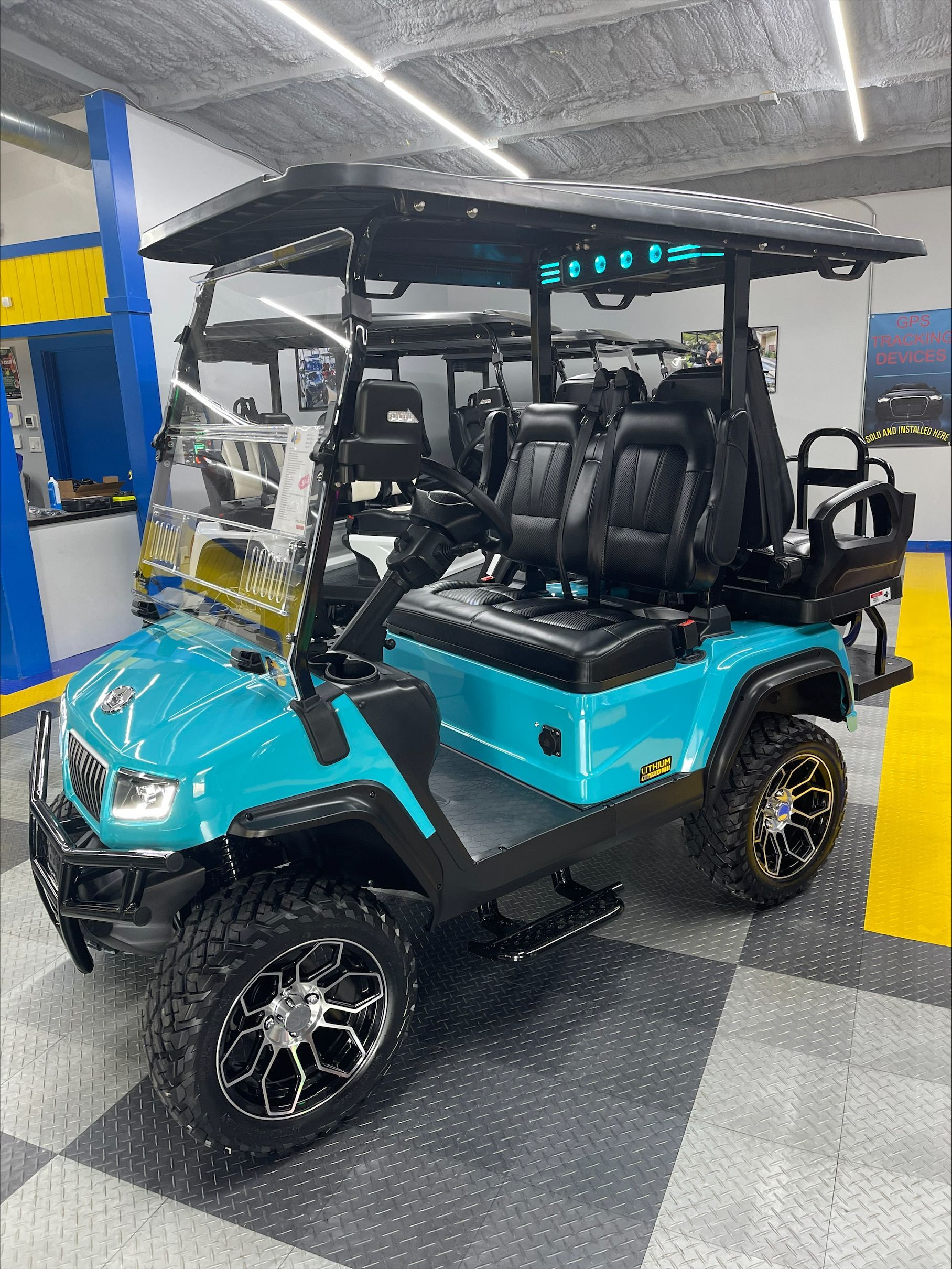 A teal golf cart with black accents, lifted tires, and four black seats parked indoors.
