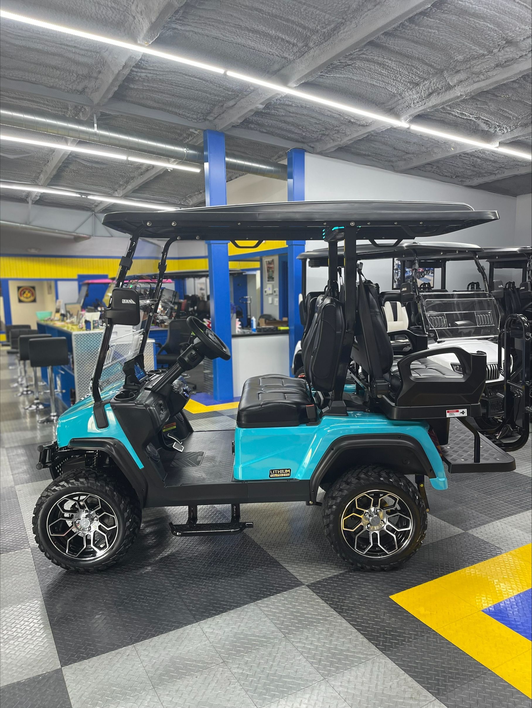 A teal golf cart with black accents and large off-road tires is parked inside a showroom.