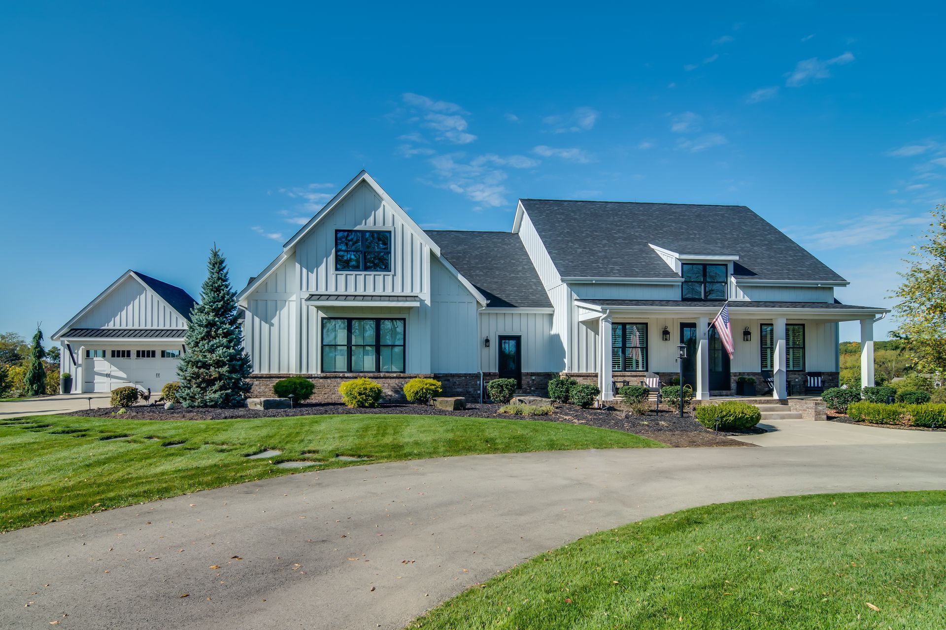 A beautiful house with black trim, a porch, and a detached garage on a grassy hill under a blue sky