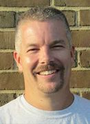 A man with a beard and mustache is smiling in front of a brick wall.