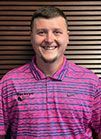 A man is wearing a pink and blue striped shirt and smiling.