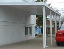 A red car is parked under a canopy in front of a white house.