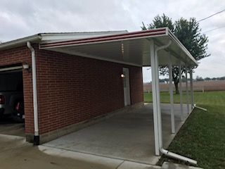 A car is parked under a canopy in front of a brick garage.