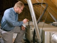 A man is kneeling down in an attic working on an air conditioner.
