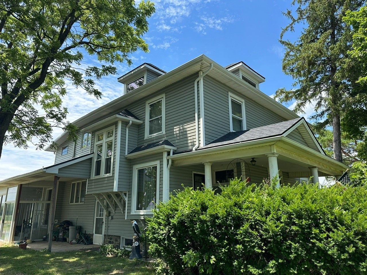 A large gray house with a porch is surrounded by trees and bushes on a sunny day.