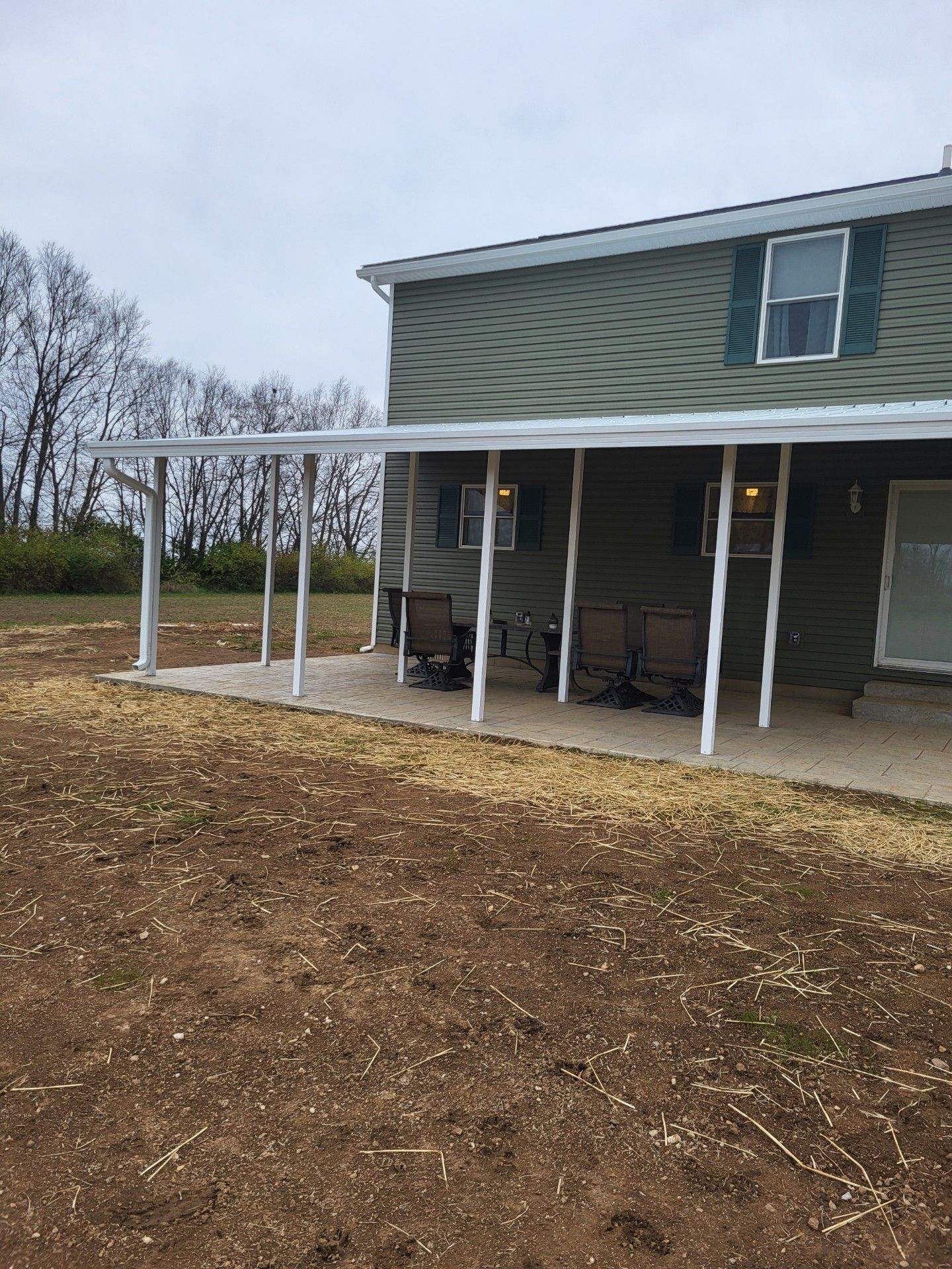 A house with a porch and chairs on it is sitting on top of a dirt field.
