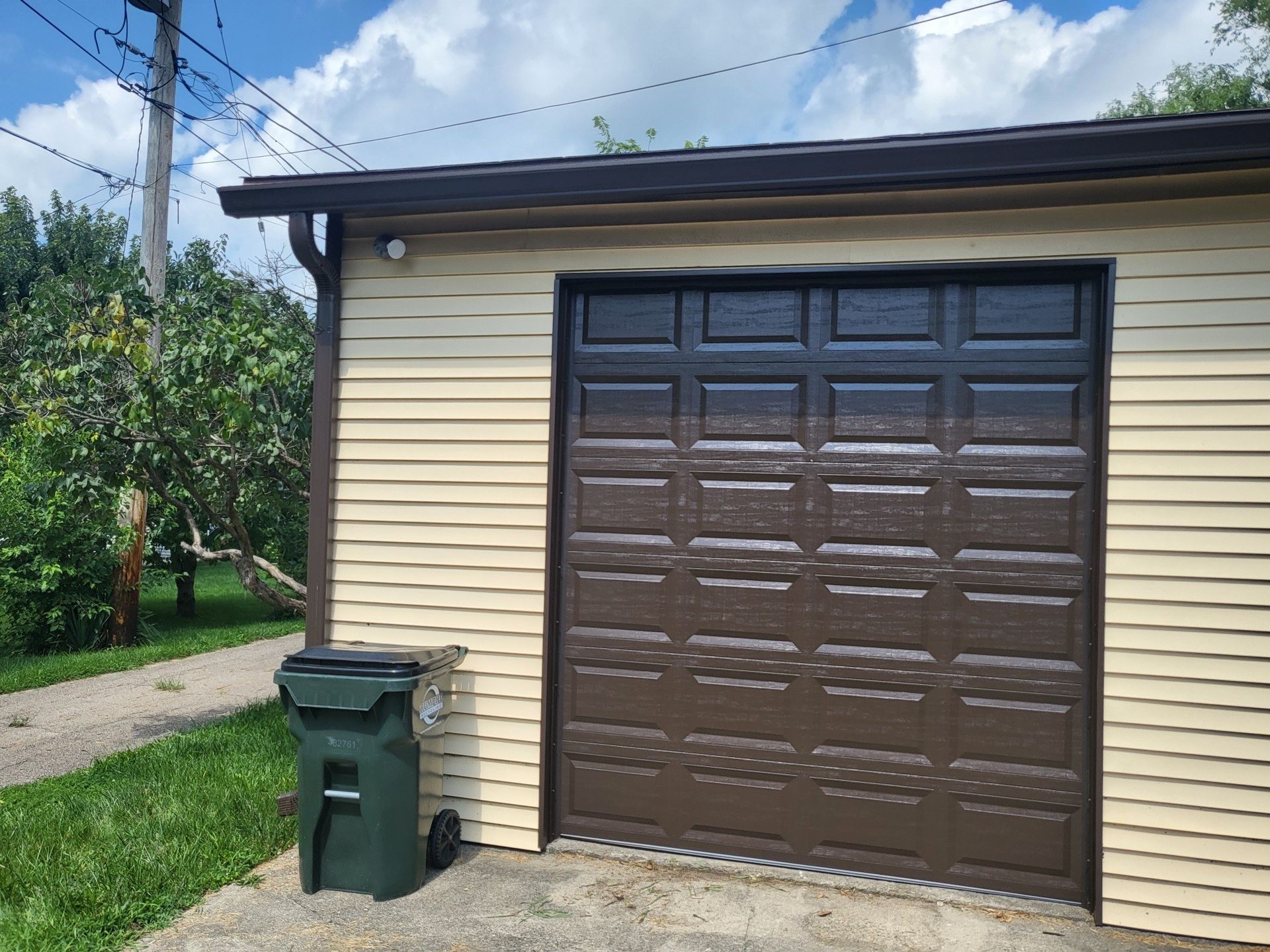 A garage with a brown garage door and a green trash can in front of it.